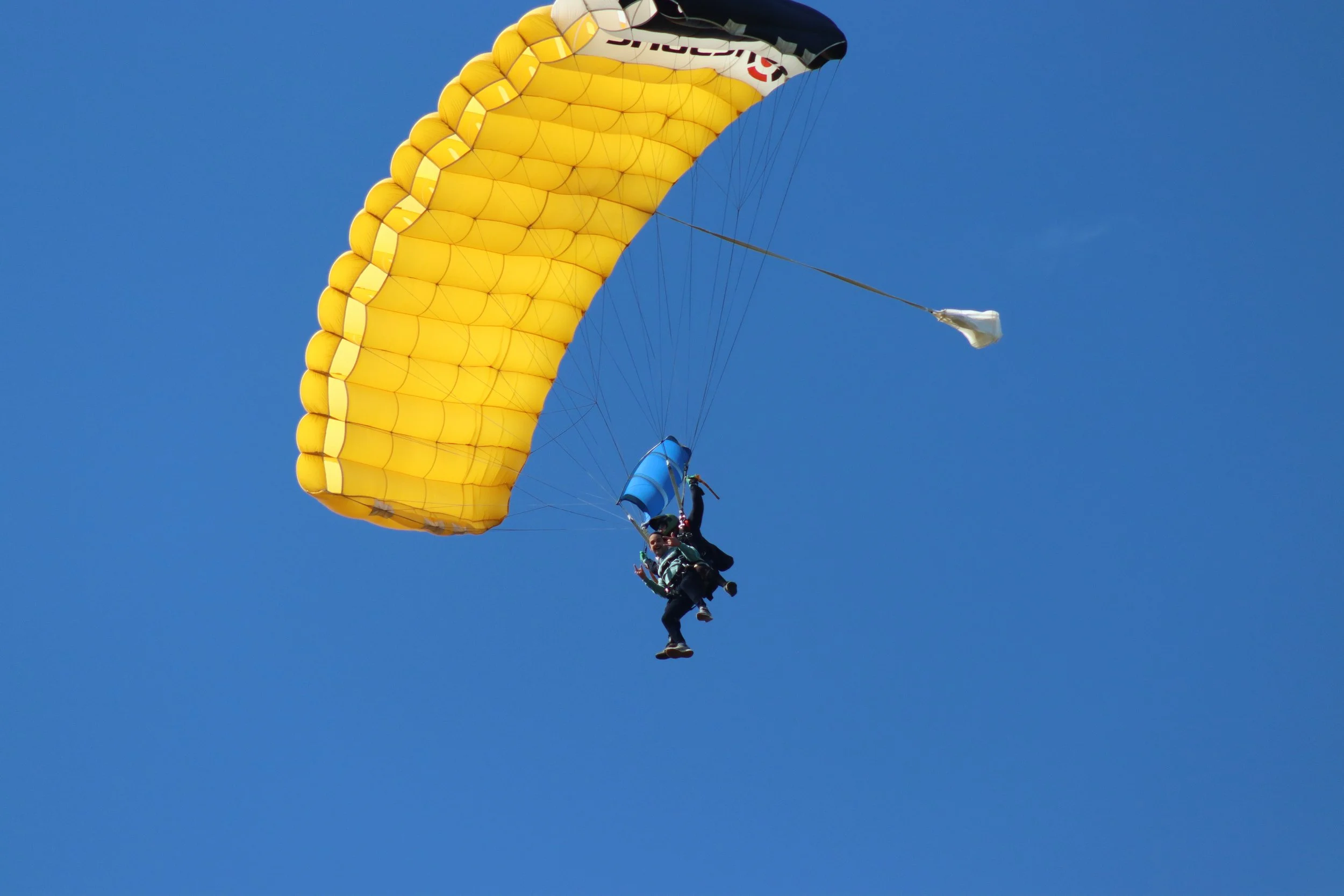 Two people with parachutes are skydiving under a bright yellow parachute against a clear blue sky.