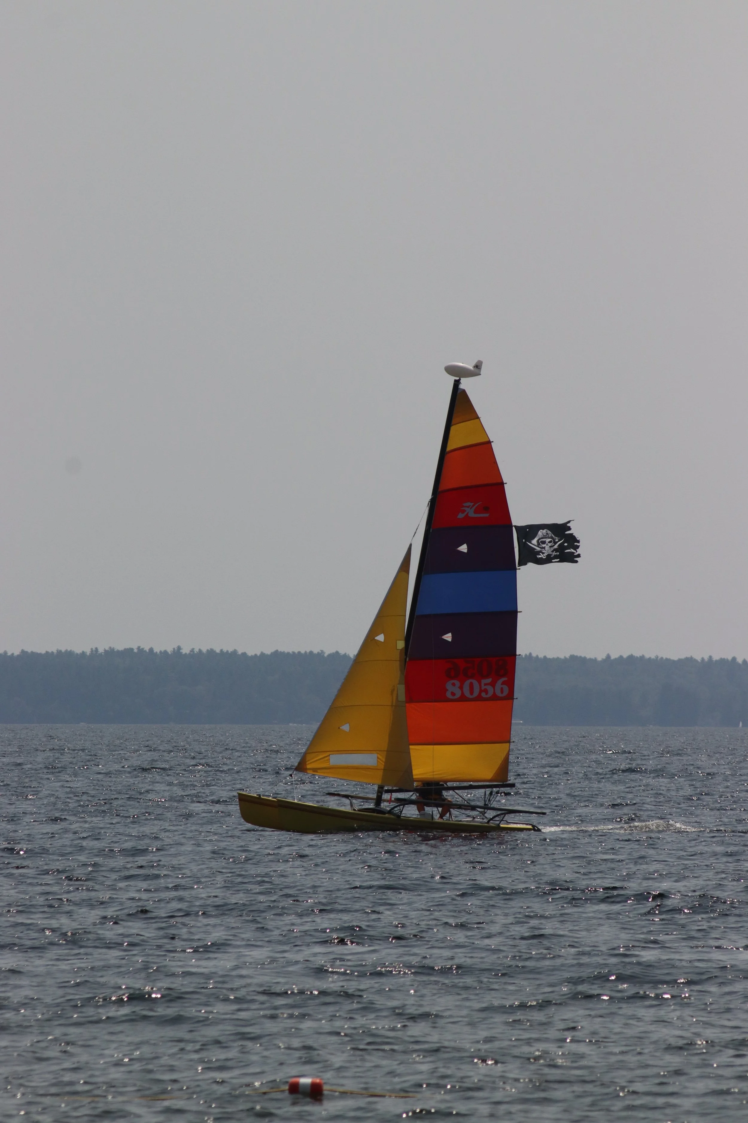 A colorful sailboat with yellow, red, blue, and black sails on a body of water, flying a pirate flag with a skull and crossbones, under a gray sky.