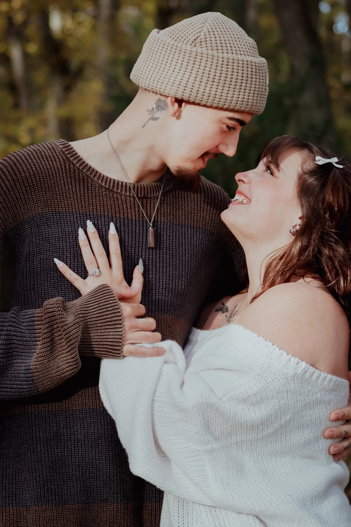 A couple is smiling and looking into each other's eyes outdoors, with trees in the background. The woman wears a white off-shoulder sweater and a hair bow, while the man wears a brown and beige striped sweater, a beige beanie, and a pendant necklace.
