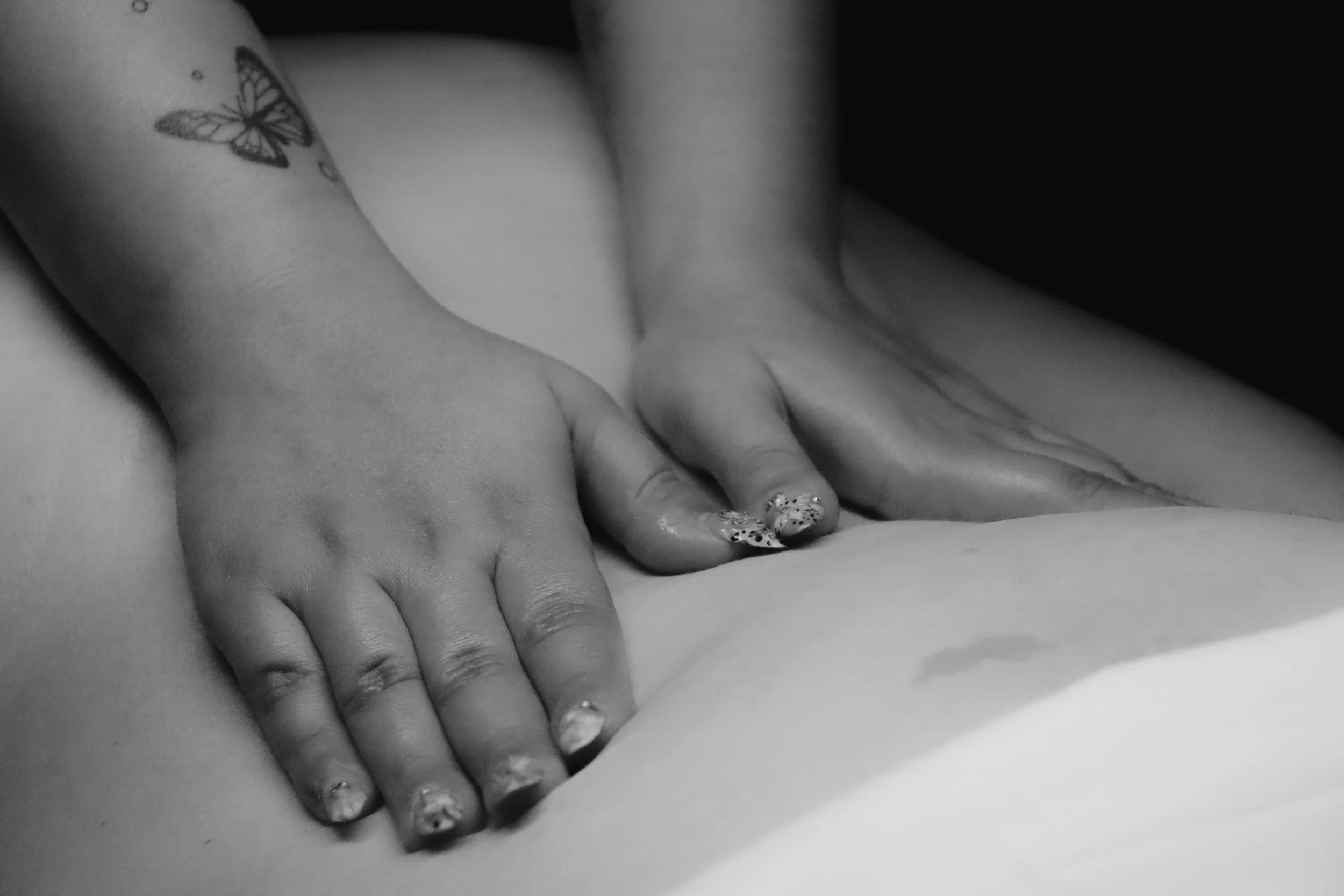 Close-up of a person's hands with a butterfly tattoo, pressing down on another person's back during a massage, in black and white.