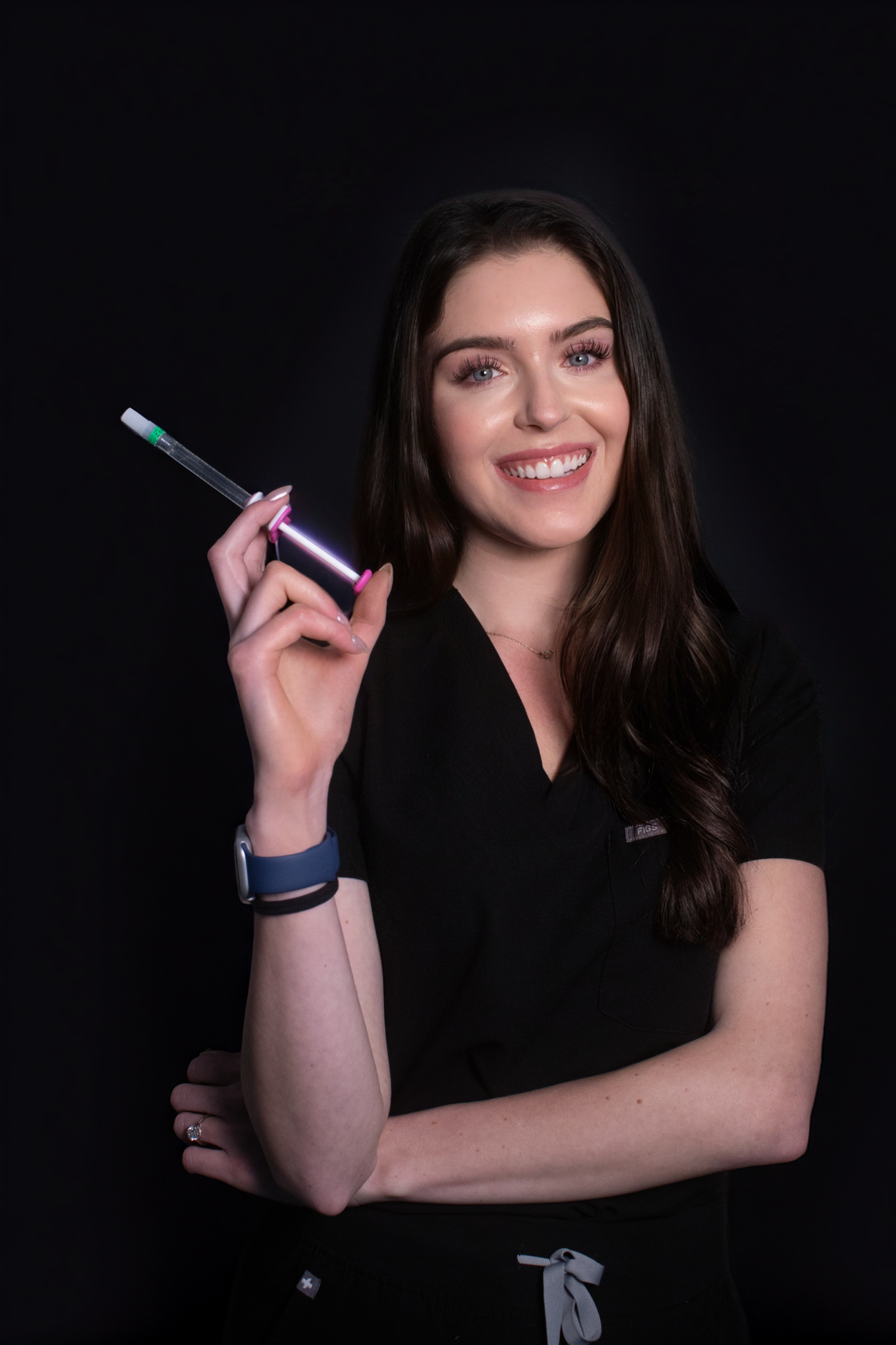 A woman with long dark hair smiling while holding a syringe, against a dark background.