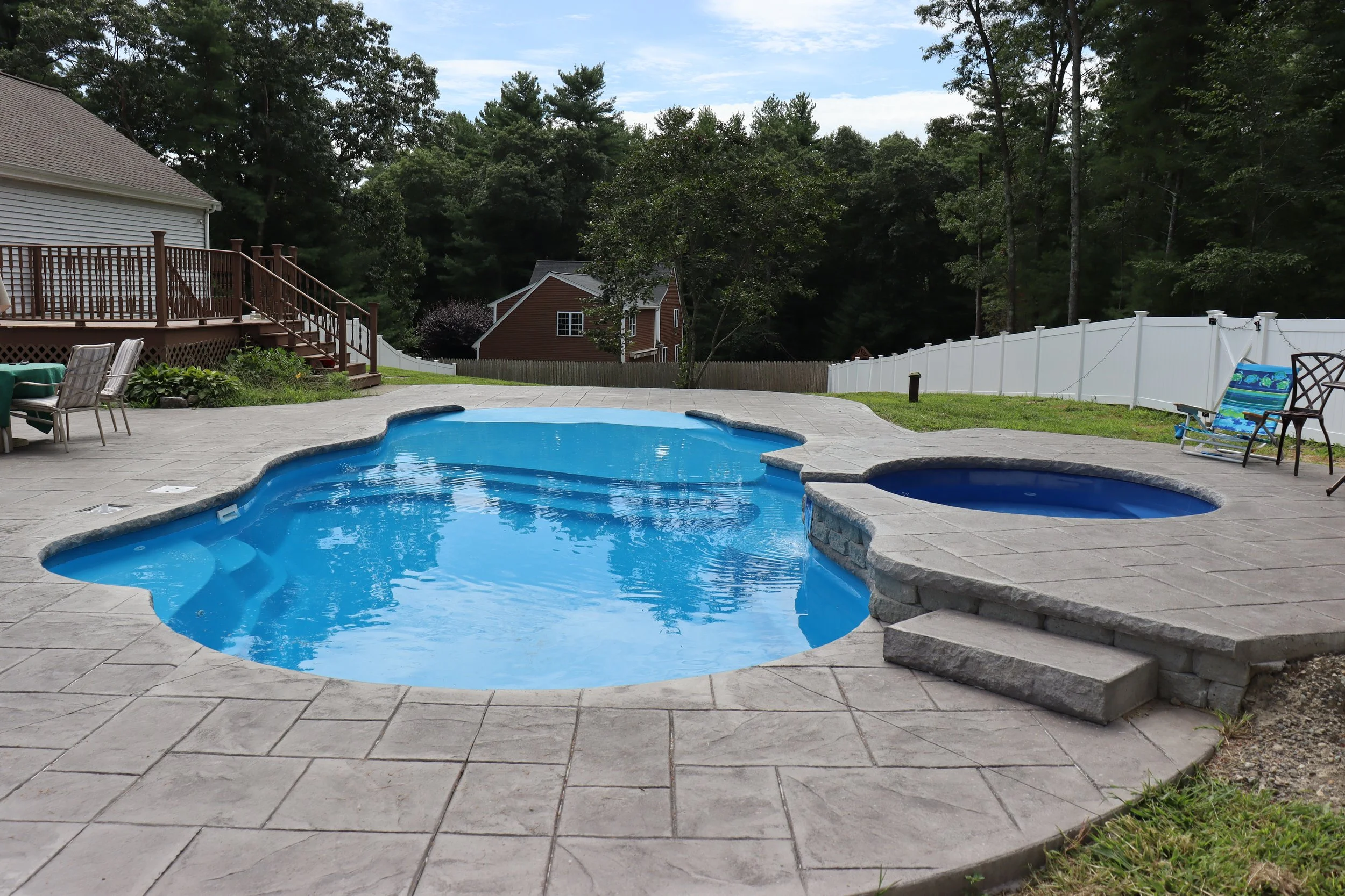 Backyard with a kidney-shaped swimming pool, hot tub, and outdoor seating surrounded by a concrete deck, wooden fence, trees, and neighboring houses.