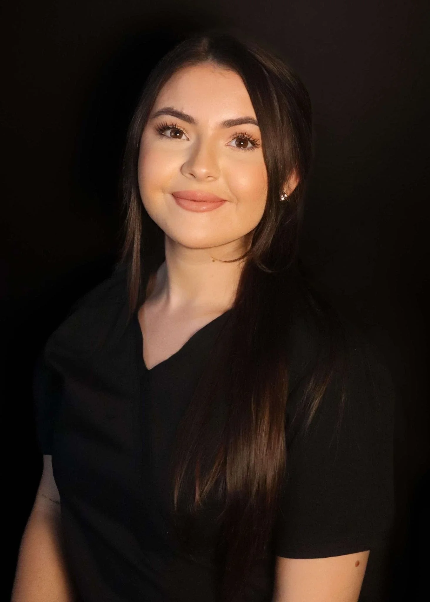 A young woman with long brown hair, wearing a black top, smiling in front of a black background.