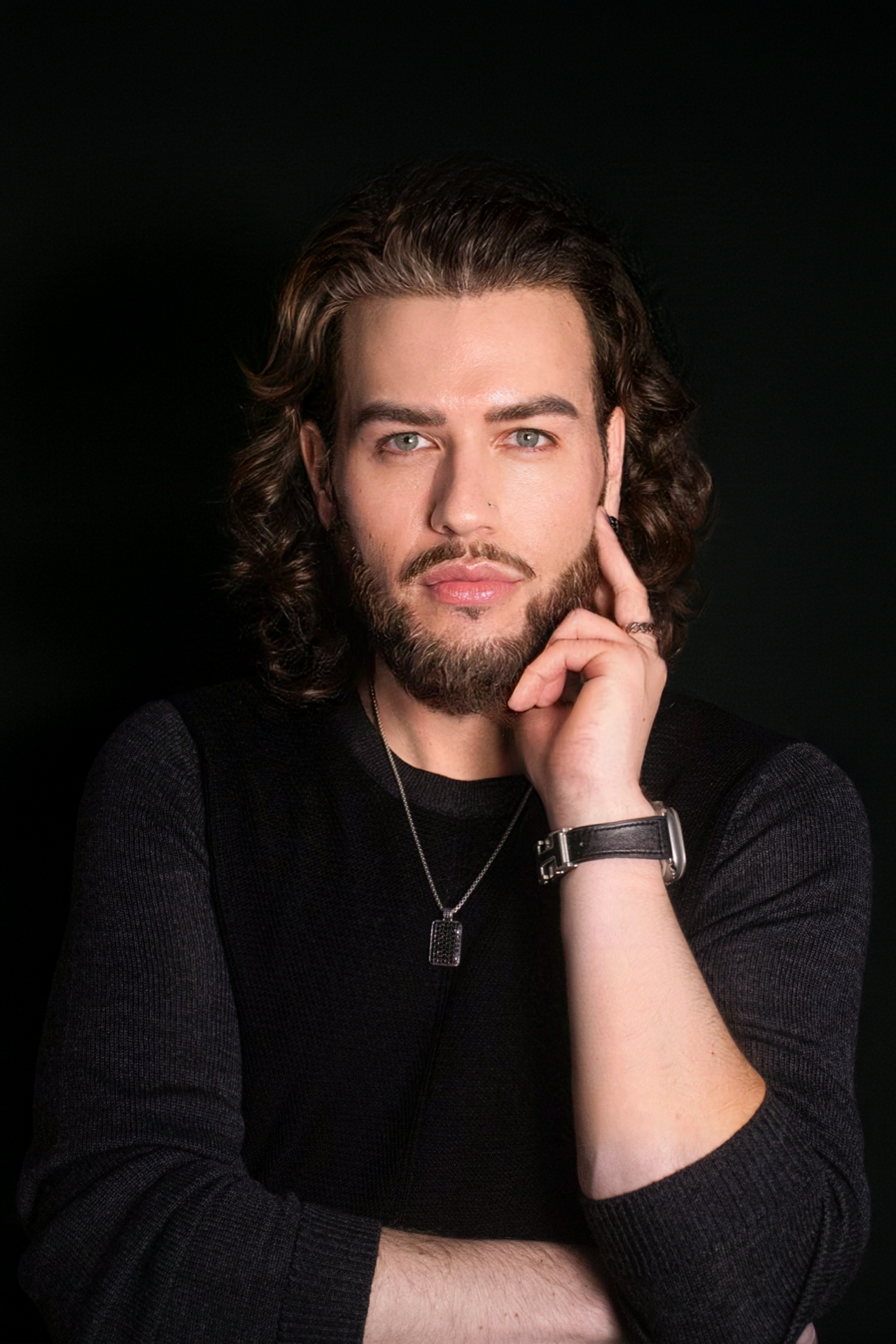 Portrait of a young man with long, curly brown hair and a beard, posing with one hand touching his face against a dark background.