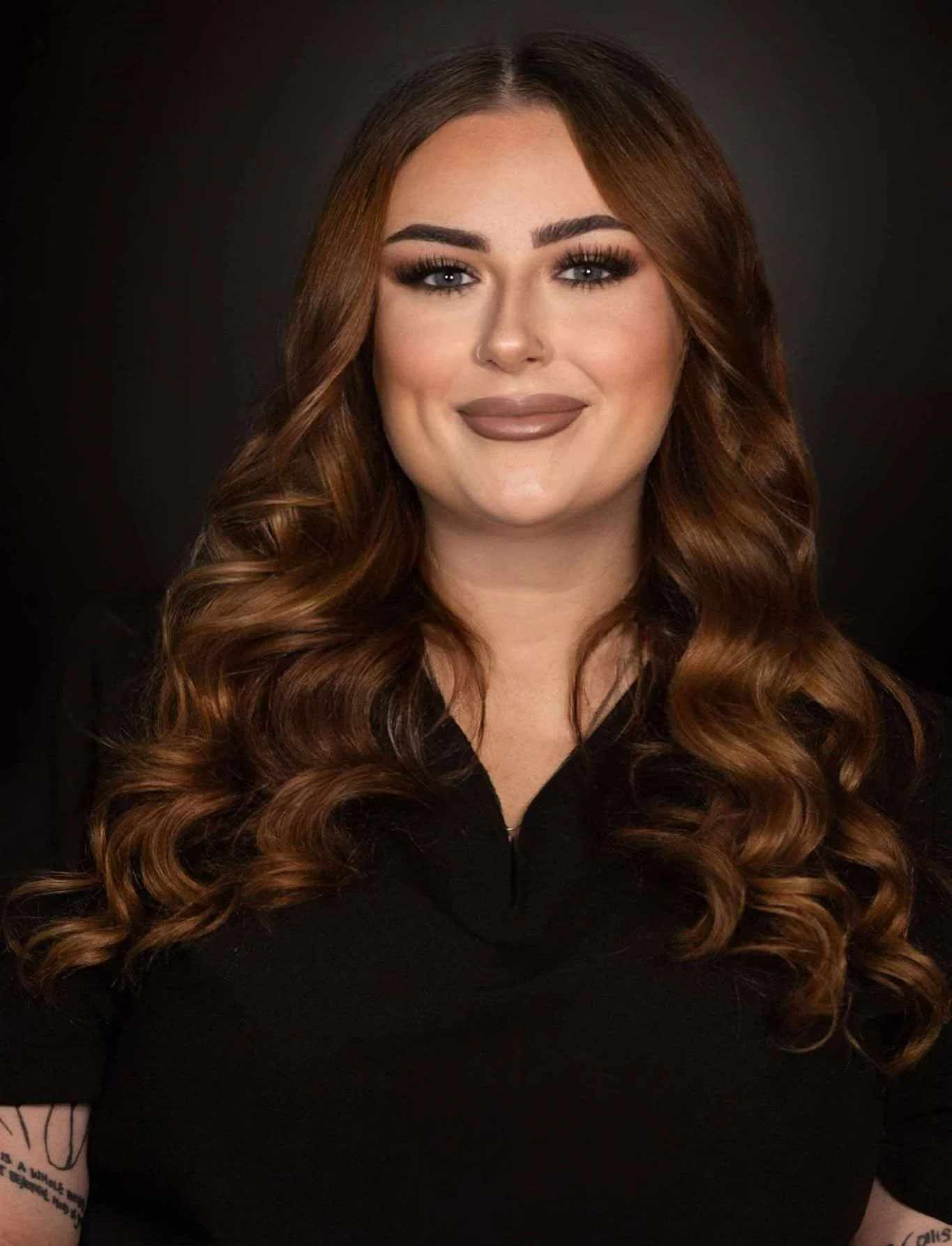 A woman with long, wavy auburn hair and makeup, wearing a black top, smiling against a dark background.
