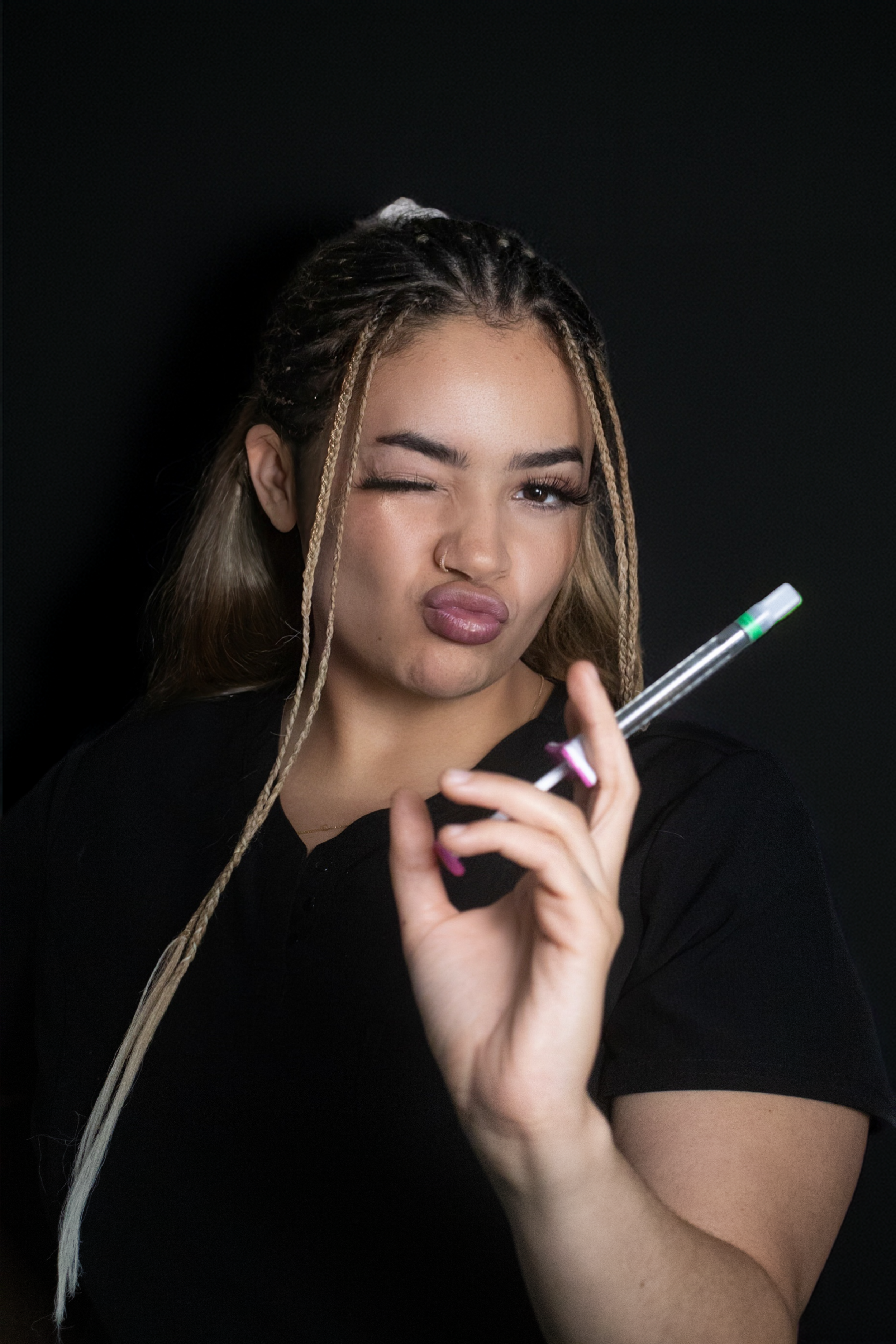 A young woman with braided hair and a nose ring holding a syringe, winking and making a playful gesture against a black background.