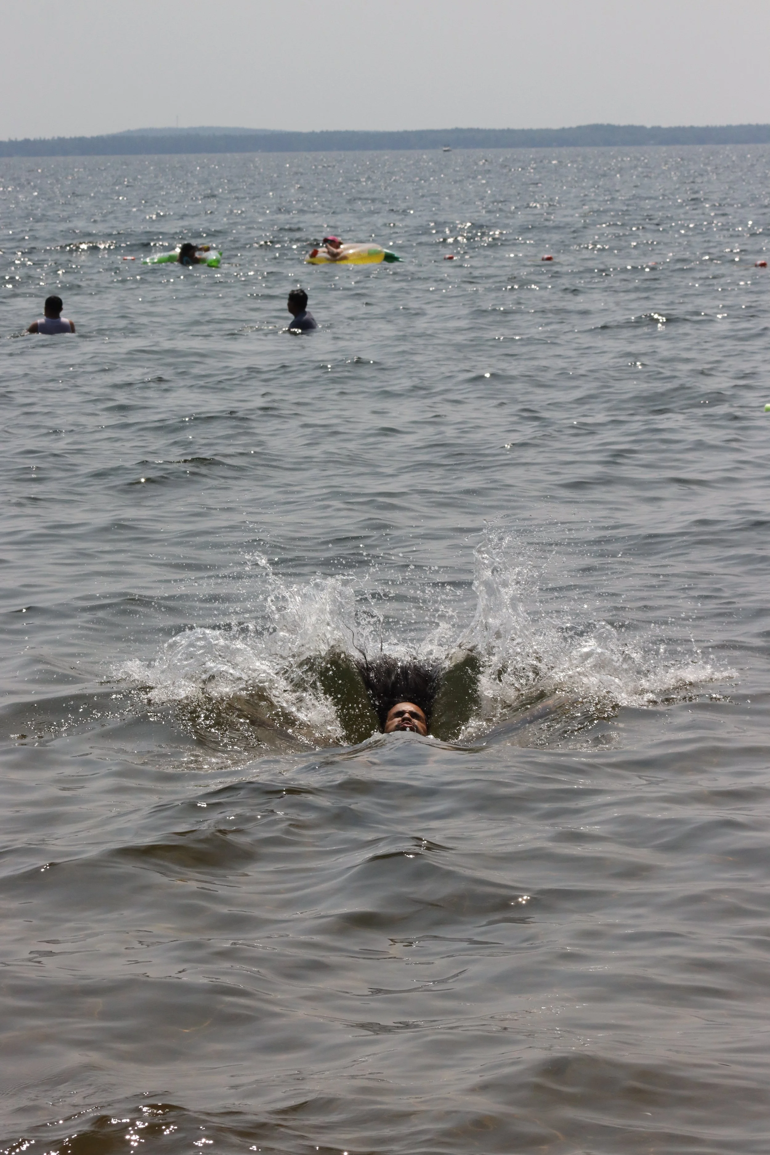 Person swimming and diving in a large body of water on a cloudy day, with other people swimming and floating with inflatable tubes in the background.