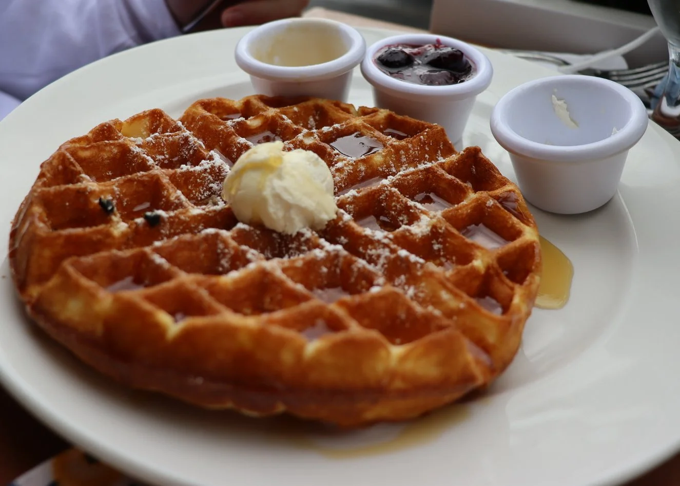 A plate with a large waffle topped with a dollop of butter and powdered sugar, served with small cups of syrup, fruit preserves, and cream.