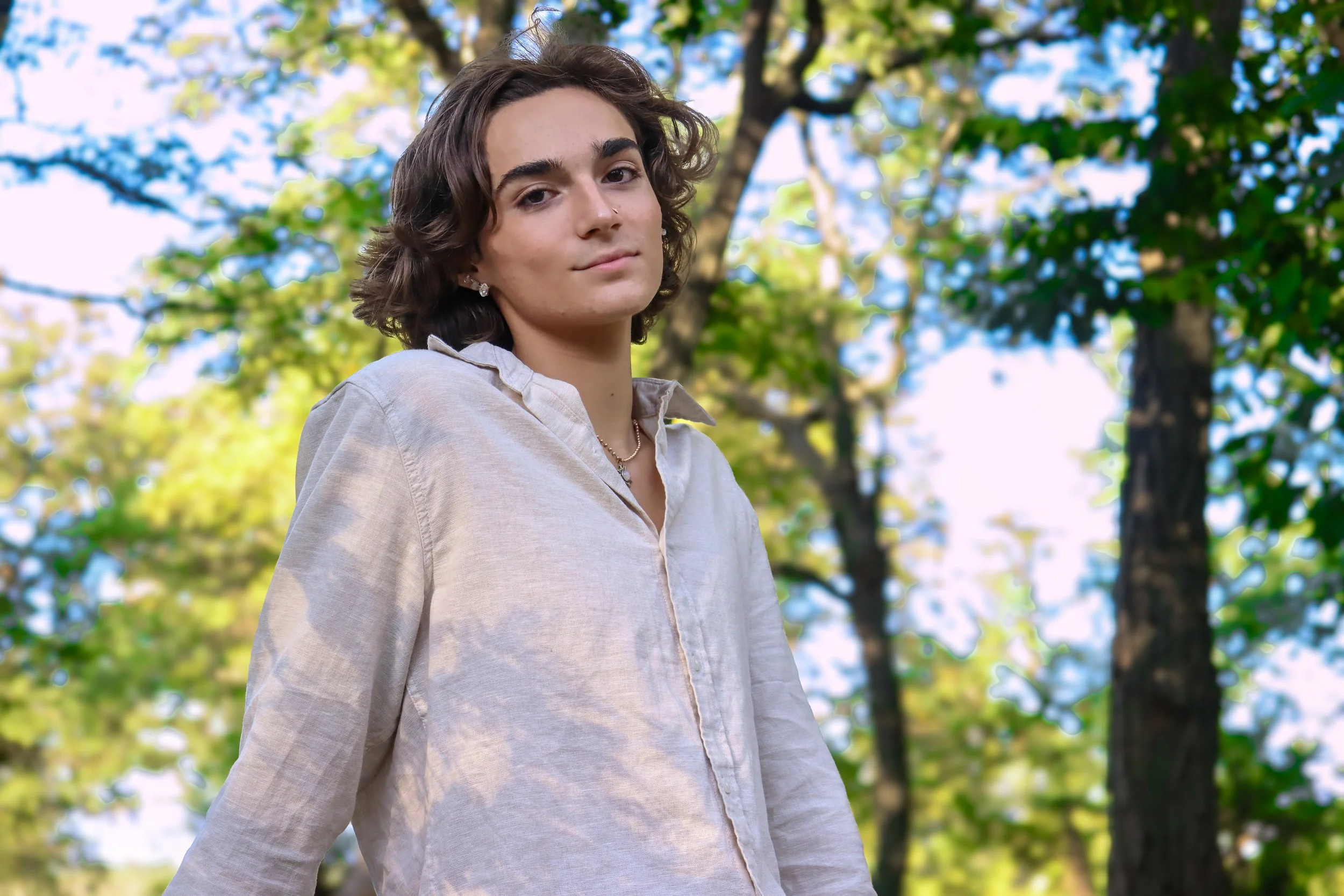 A young person with shoulder-length brown hair standing outdoors in a park with green trees and blue sky in the background, wearing a light-colored button-up shirt and looking at the camera.