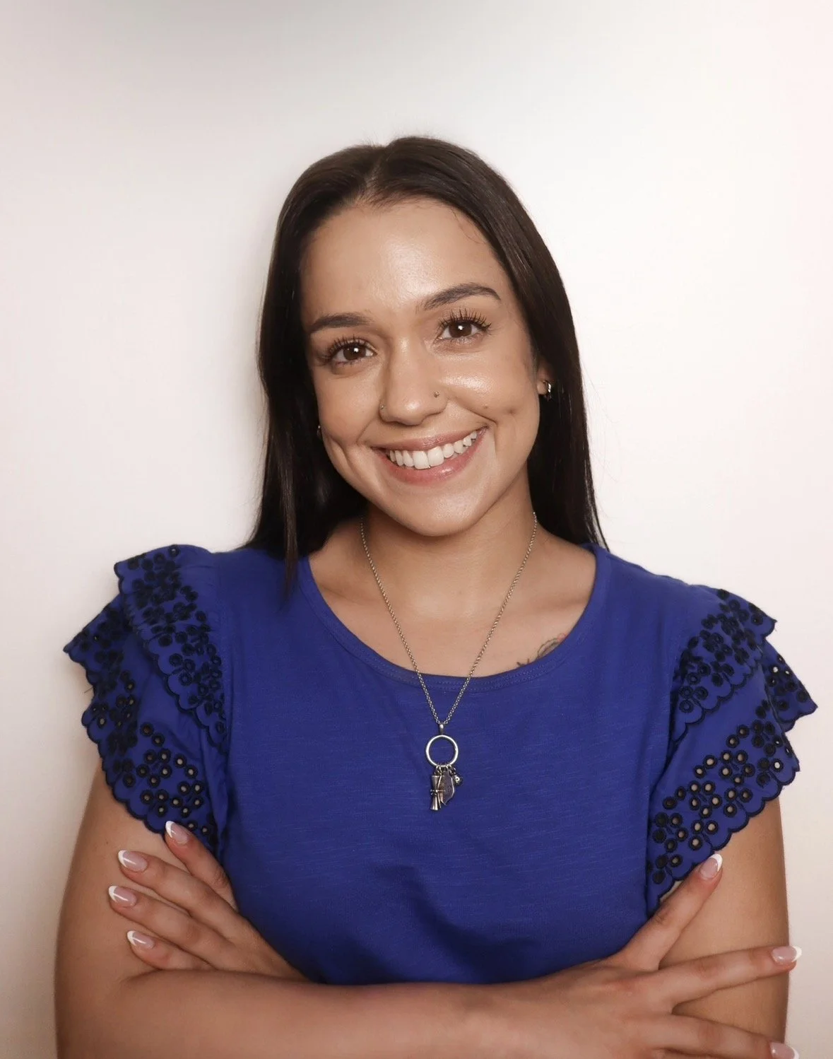 Smiling young woman with long dark hair, wearing a blue top with decorative sleeves and a necklace with a ring and charms, standing against a plain white wall.