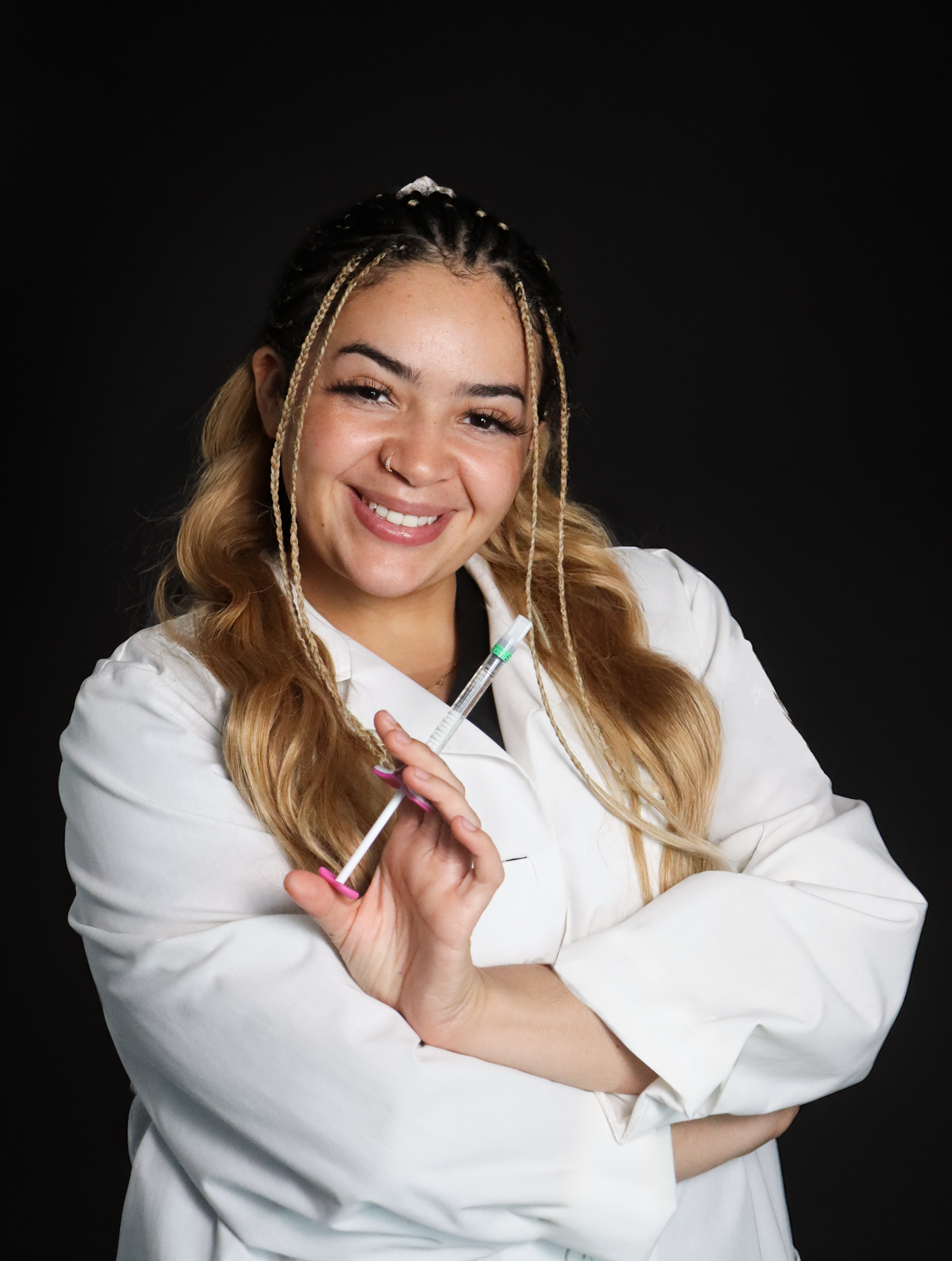 Woman smiling in a white lab coat holding a syringe against a black background.