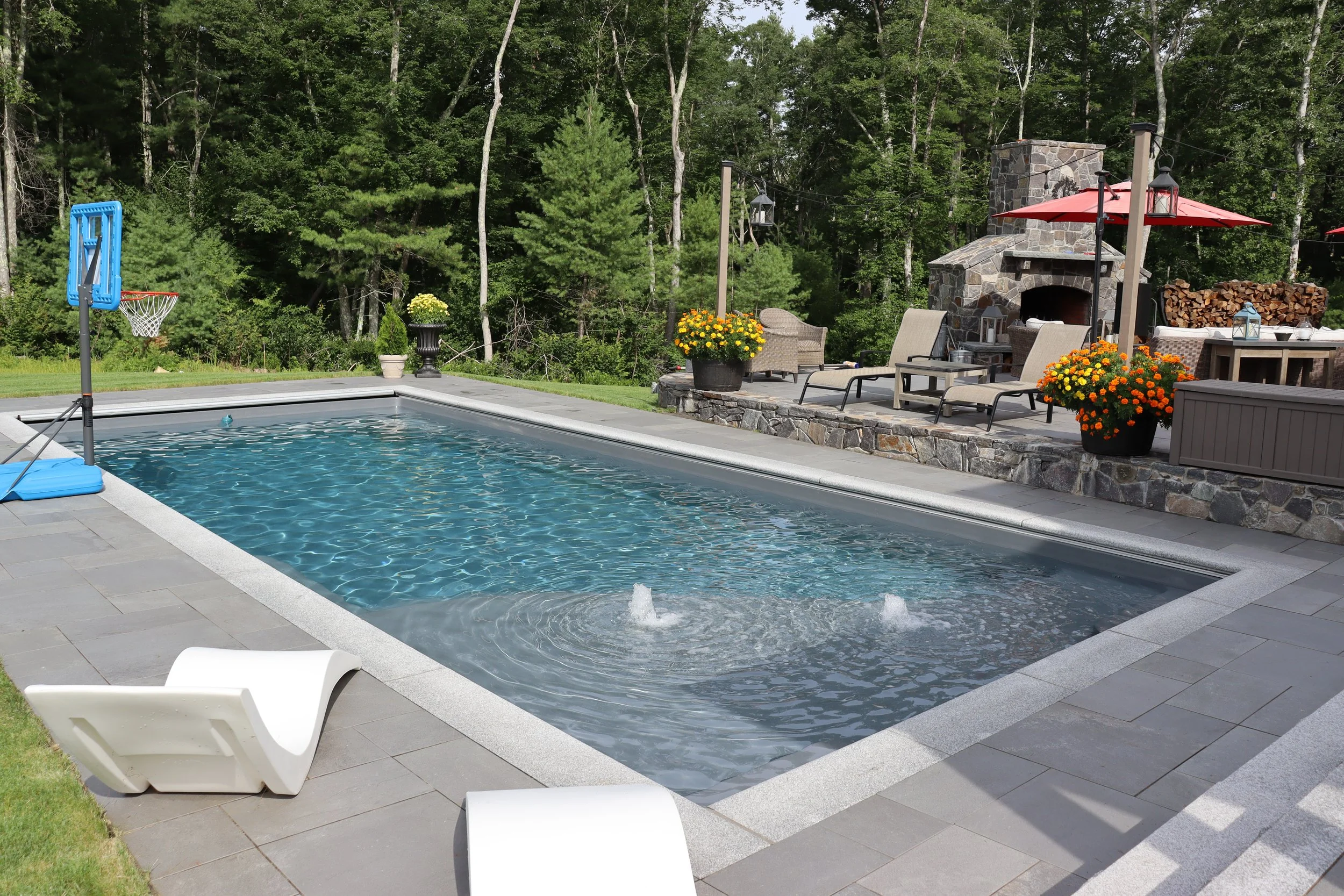 Backyard swimming pool with a fountain feature, surrounded by stone patio and outdoor furniture, including lounge chairs, umbrellas, and a fireplace, with lush green trees in the background.