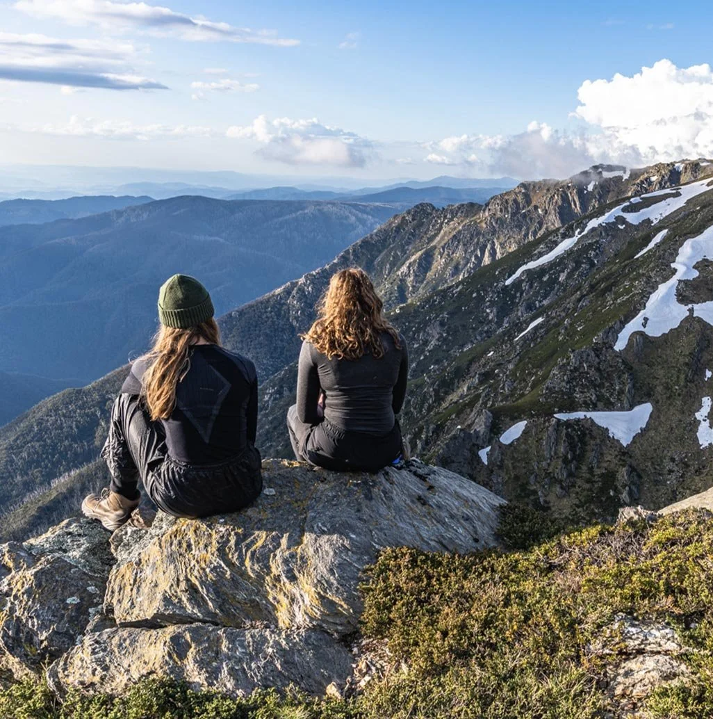 Two individuals sit atop a boulder, overlooking the expansive vista of the Snowy Mountains in Kosciuszko National Park, NSW, Australia.