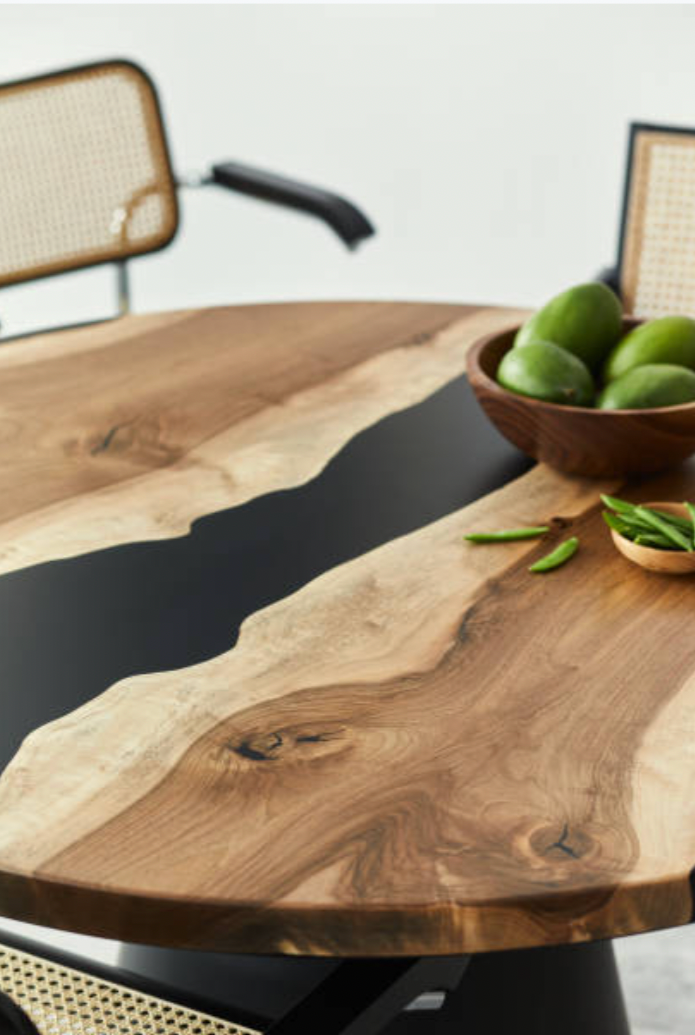 A wooden dining table with a black resin inlay, a wooden bowl of green apples, and green leaves, with woven cane chairs in the background.
