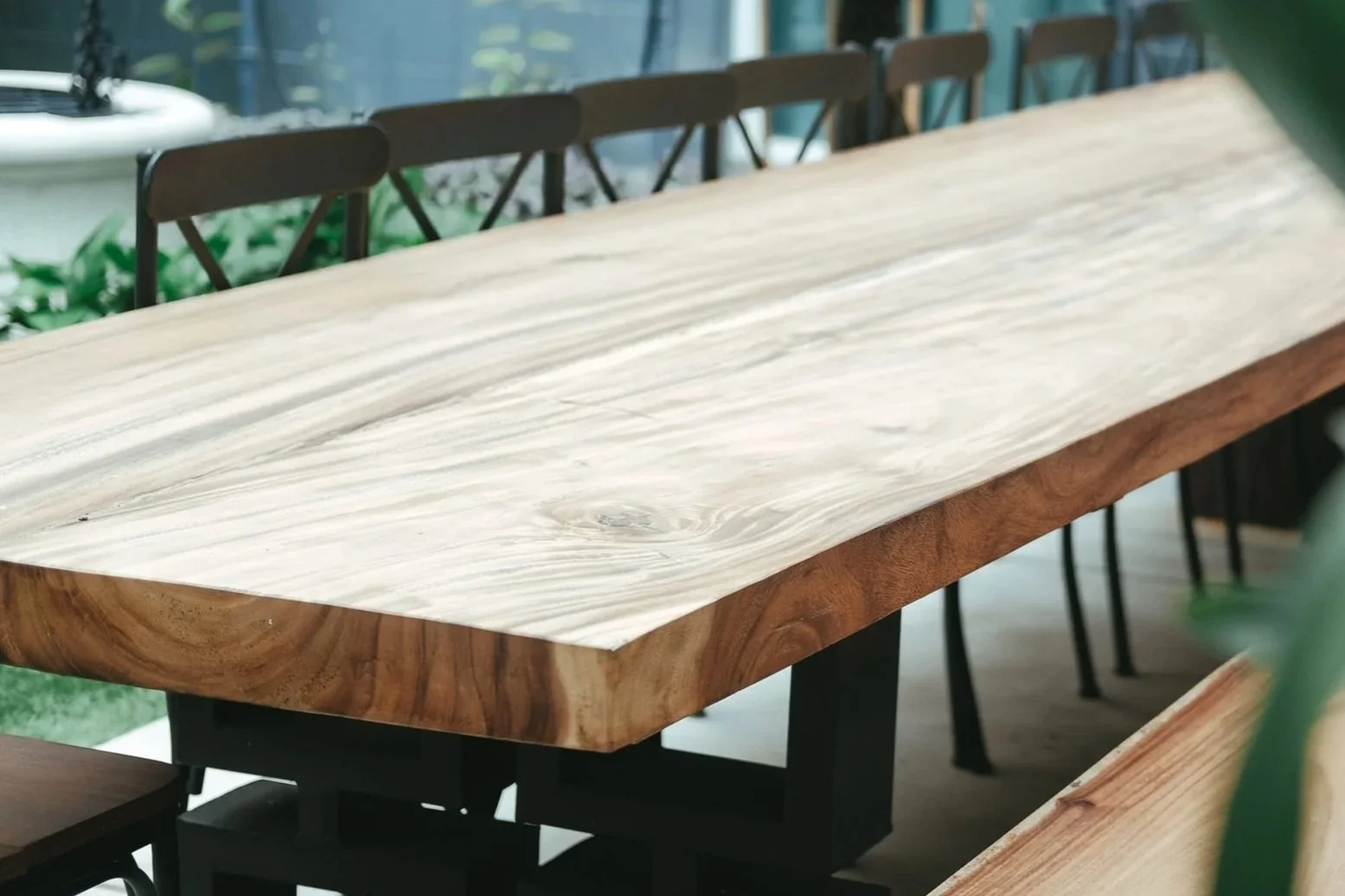 Wooden dining table with a natural edge, surrounded by black chairs, in a room with large windows and green plants in the background.