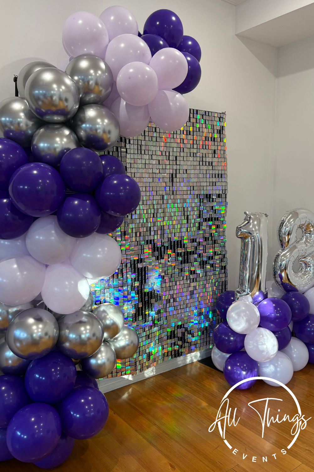 Decorative party balloons in purple, white, and silver, arranged around a shimmering mosaic backdrop with rainbow reflections, along with large silver number balloons displaying '12'.