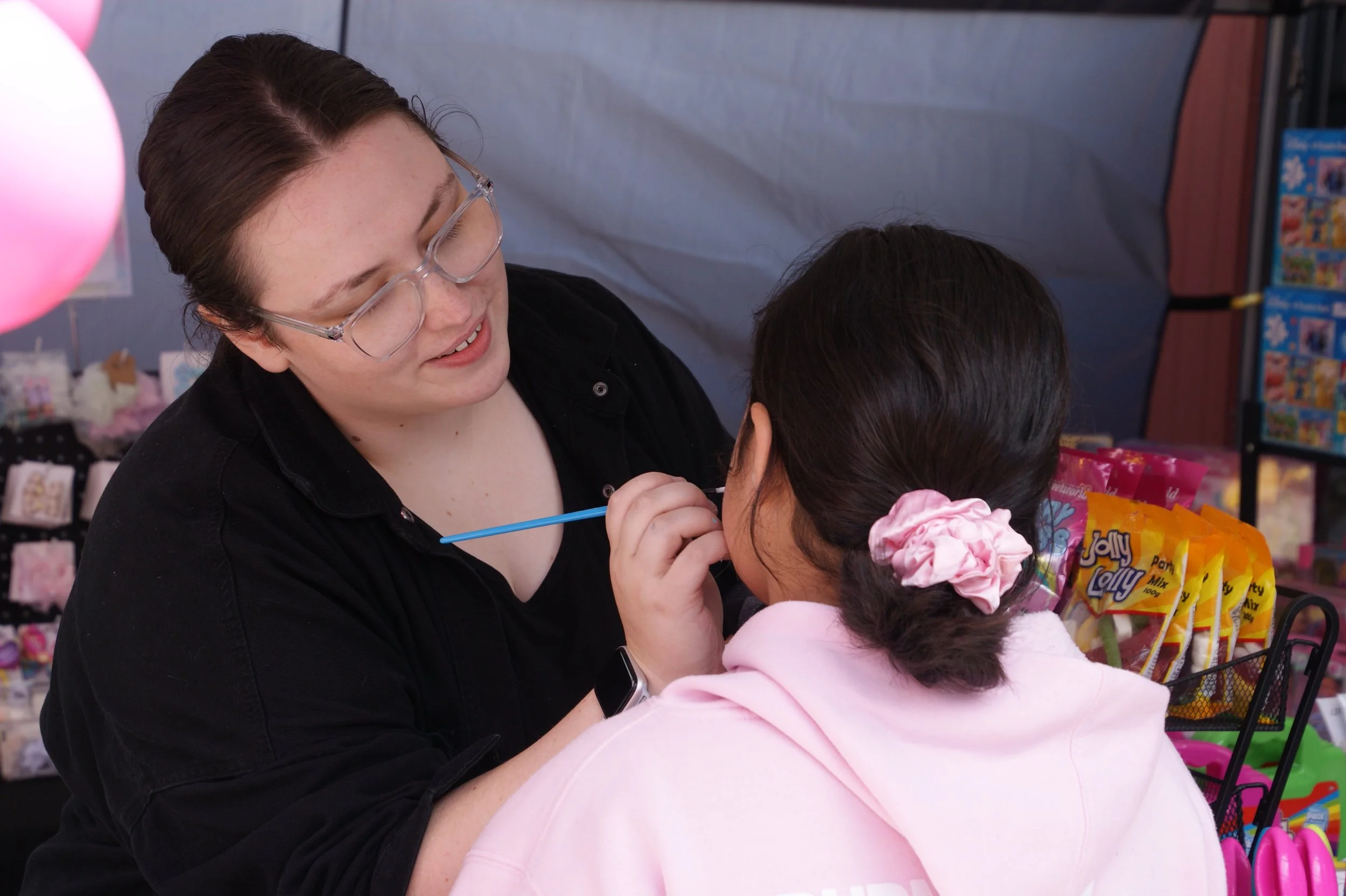 A woman getting a face painting at a birthday party, with the child wearing a pink hoodie and hair scrunchie.