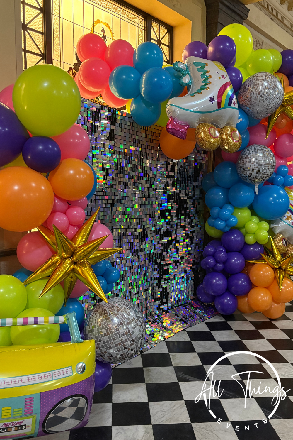 Colorful balloon arrangement with a unicorn-shaped foil balloon, disco balls, and a shiny sequin backdrop at a party or celebration.