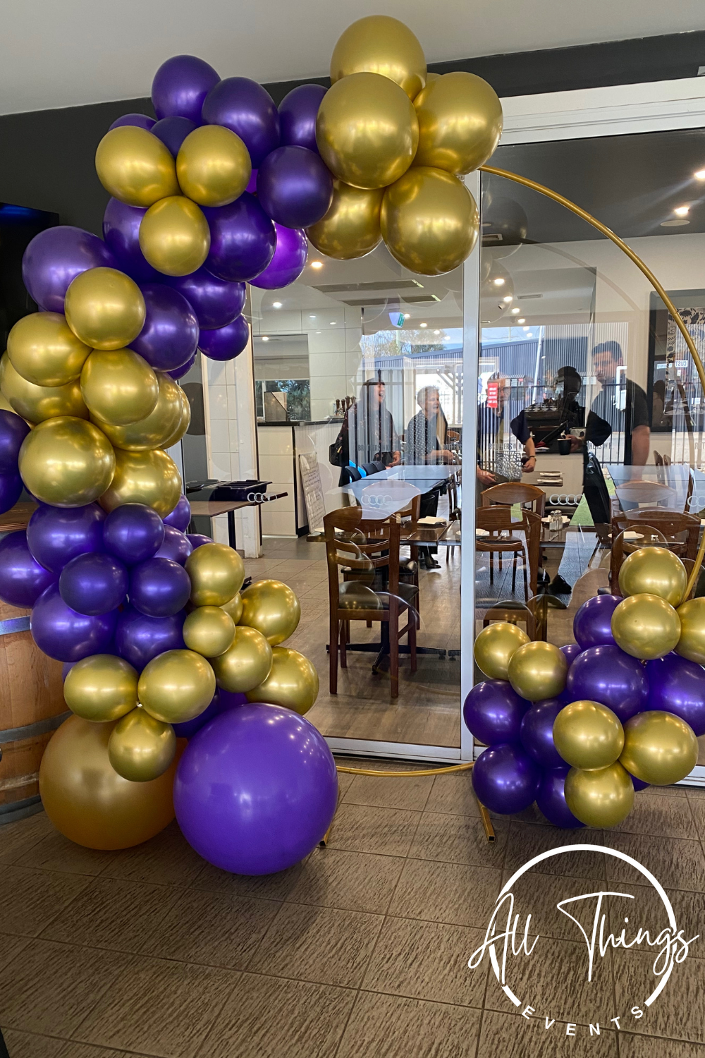 A decorative balloon arrangement with purple and gold balloons, set in front of a glass window of a restaurant or cafe, partially visible inside with wooden chairs and tables, and people dining and staff working inside.