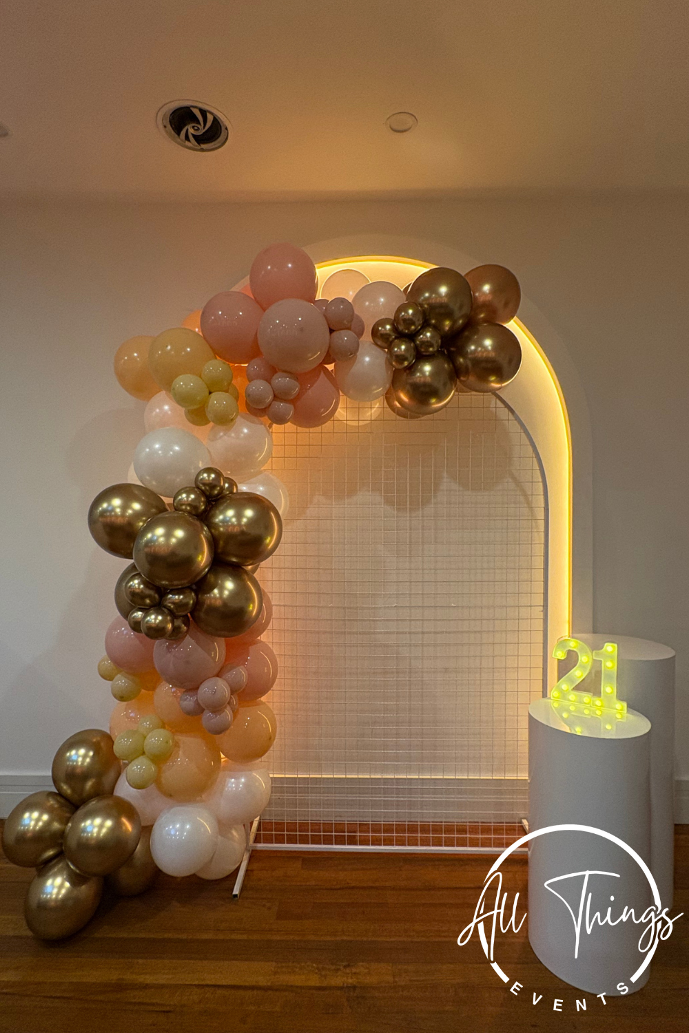 Balloon decoration with peach, white, yellow, and gold balloons arranged in an arch around a gold-lit arched backdrop, with a lit 21 sign on a white pedestal