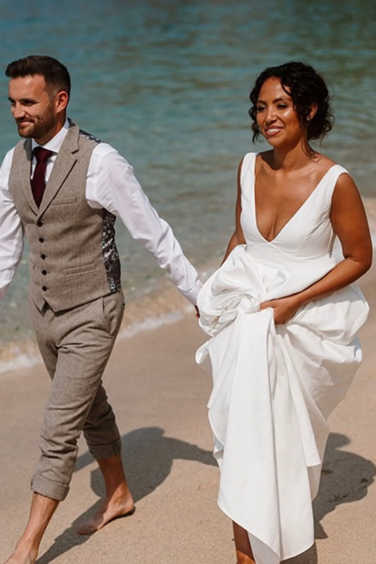 A couple walking hand in hand along the beach, the woman in a white dress and the man in a beige vest and pants, both barefoot with the ocean in the background.