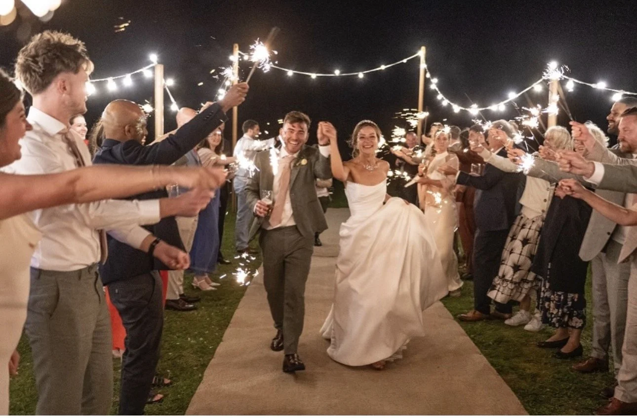 A newlywed couple walking hand in hand at night, surrounded by friends and family holding sparklers in a celebration.