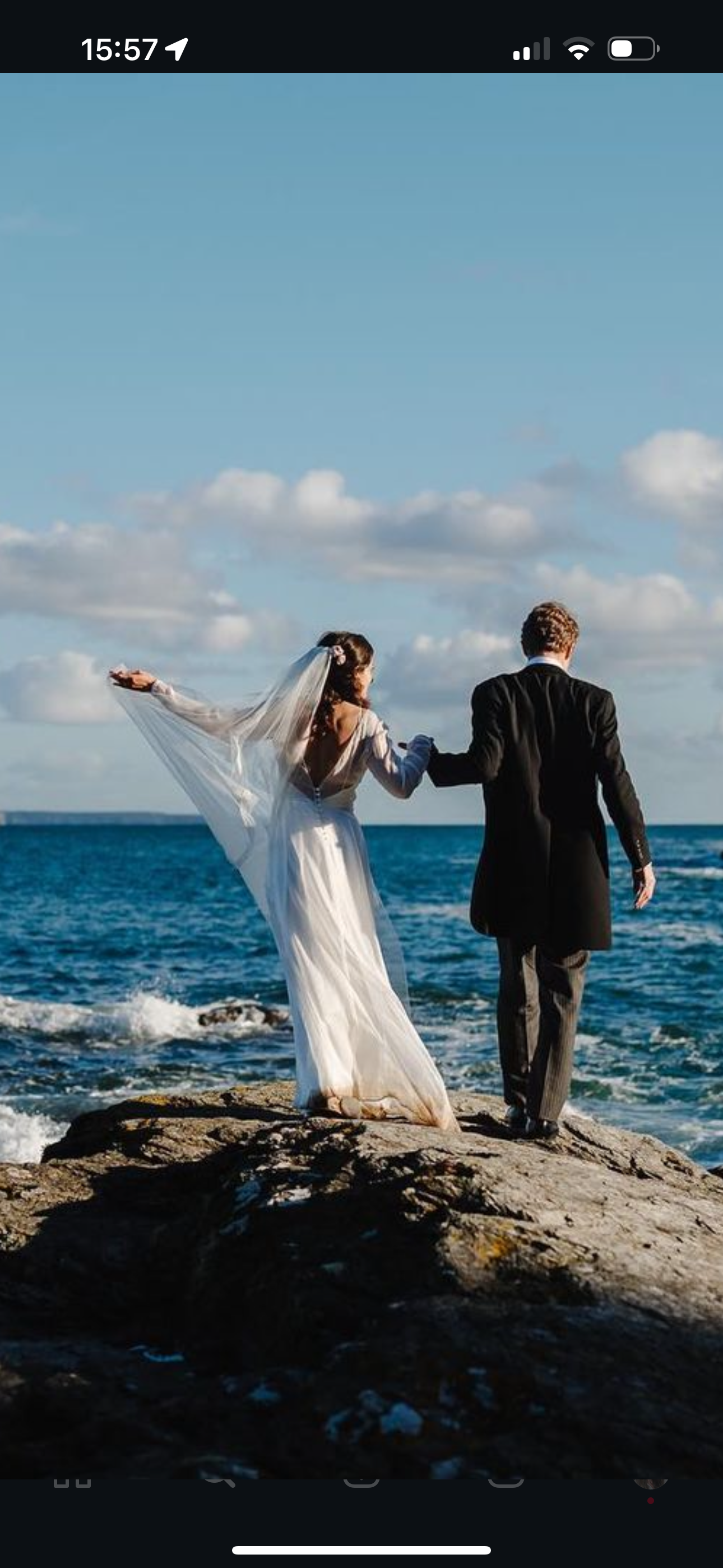 A bride and groom holding hands on rocks by the ocean, with a blue sky and clouds in the background. The bride is in a white wedding dress with a veil, and the groom is in a black tuxedo.