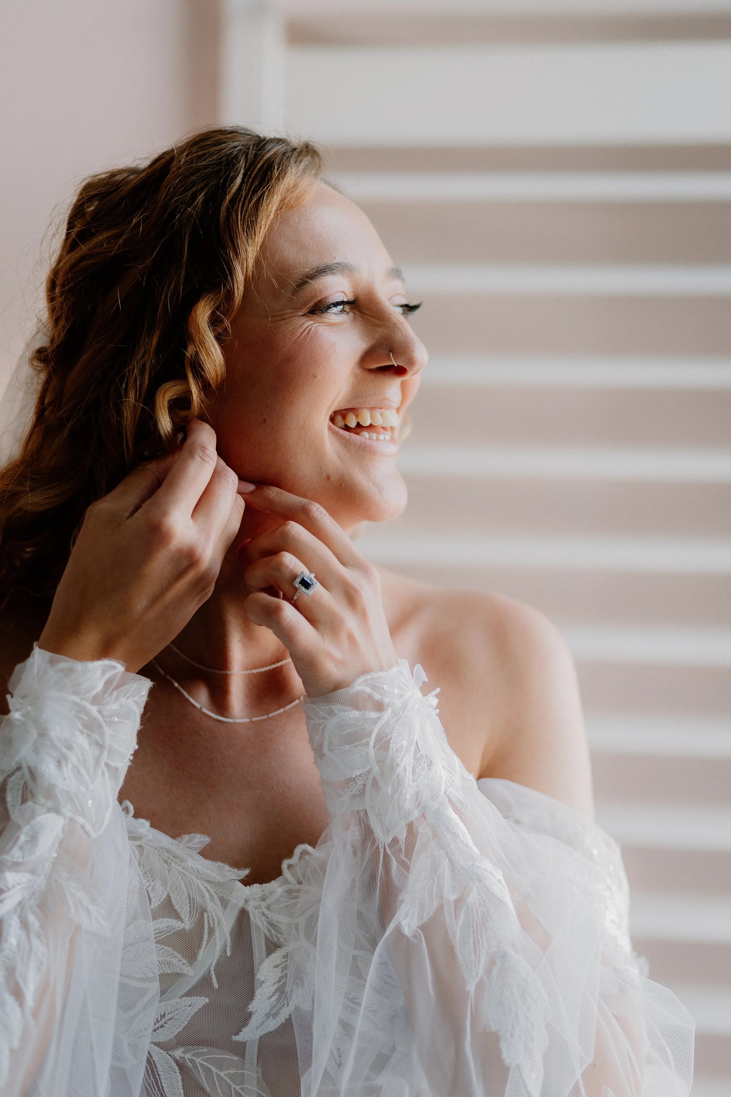A woman with light skin and wavy auburn hair smiling and looking to the right, wearing a white lace dress and a ring, with a white background with horizontal lines.