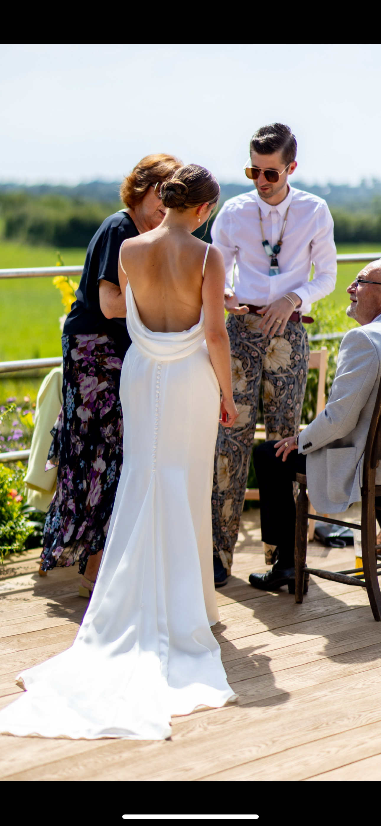 Bride in a white dress with back exposed, three people around her during outdoor wedding ceremony, lush green landscape in background.