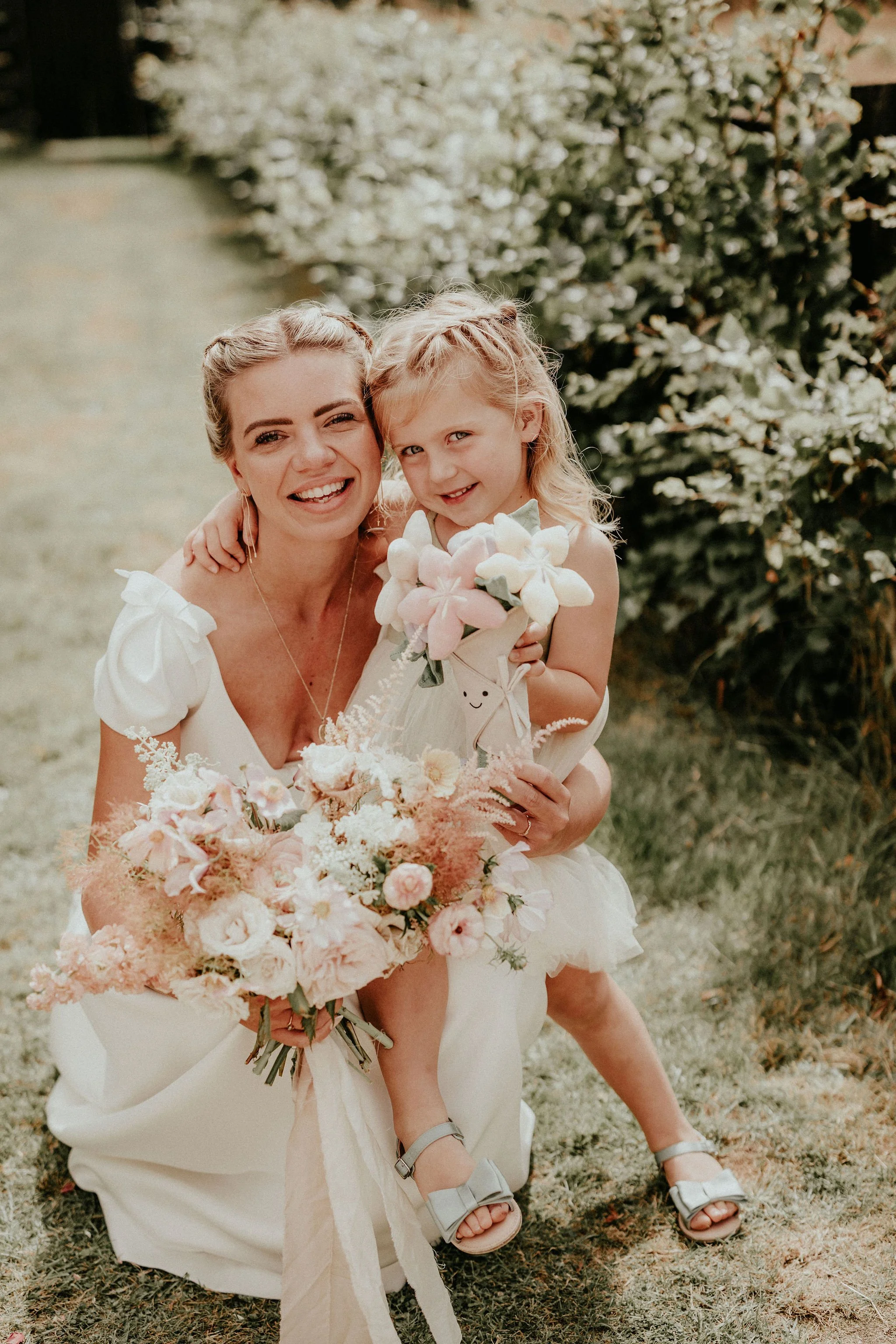 A woman and a young girl smiling outdoors, holding bouquets of flowers in a garden setting with green foliage in the background.