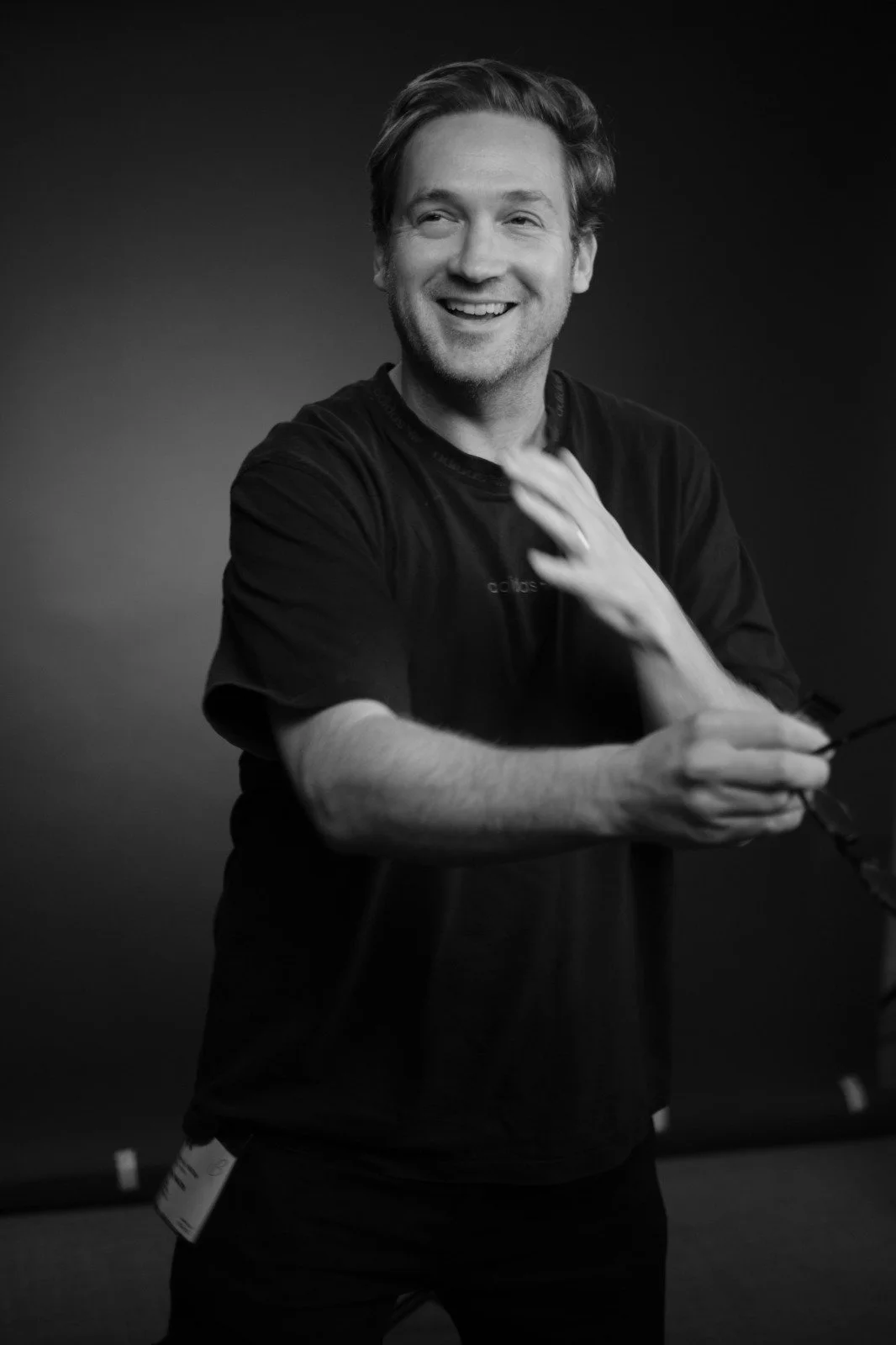 Black and white photo of a smiling man in casual clothes, holding sunglasses and adjusting his shirt.