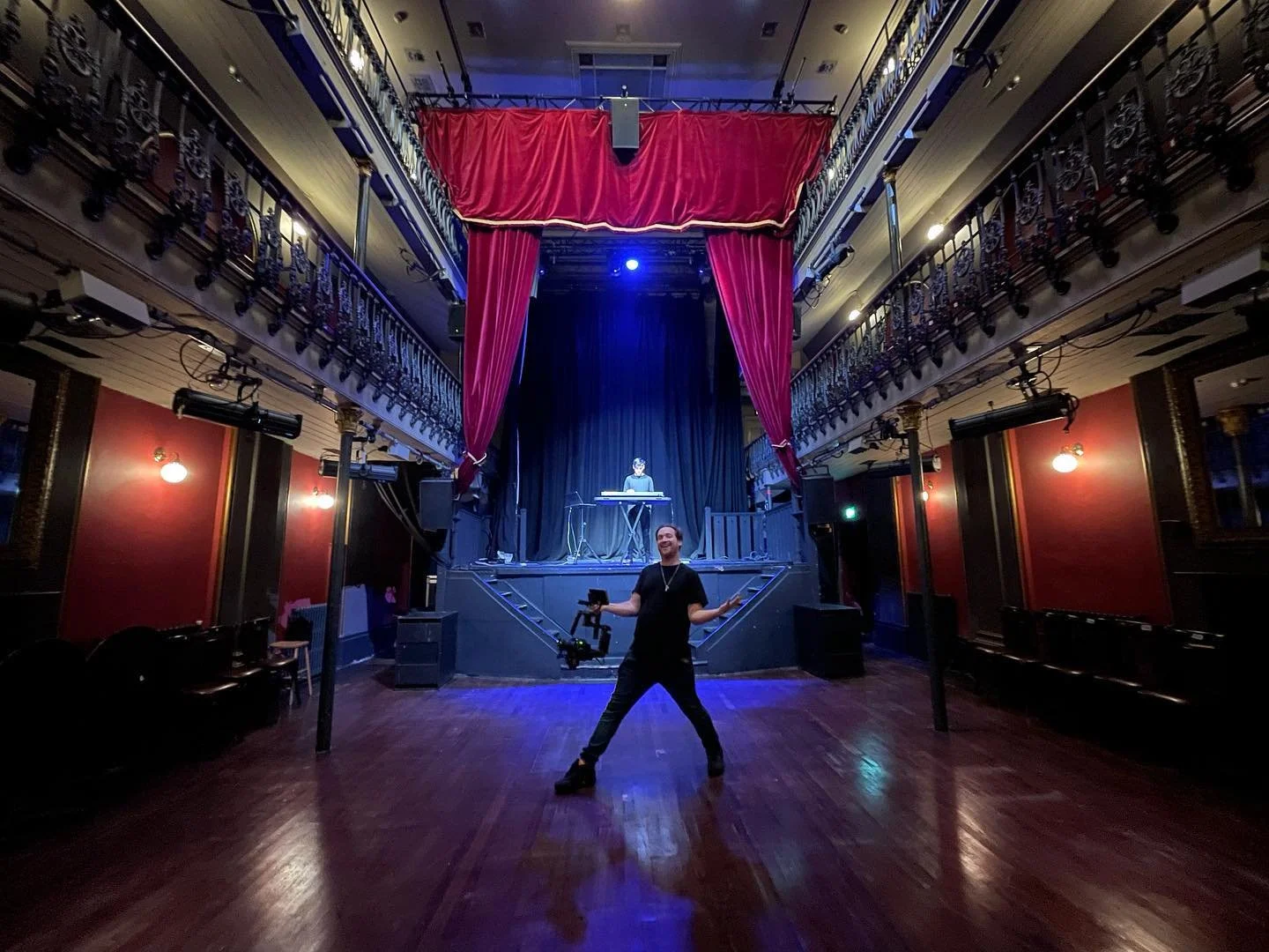 Person standing on a stage with a camera stabilizer, inside a theatre or performance venue with red curtains, a musician playing synthesizer in the background, and ornate railings on the balconies.