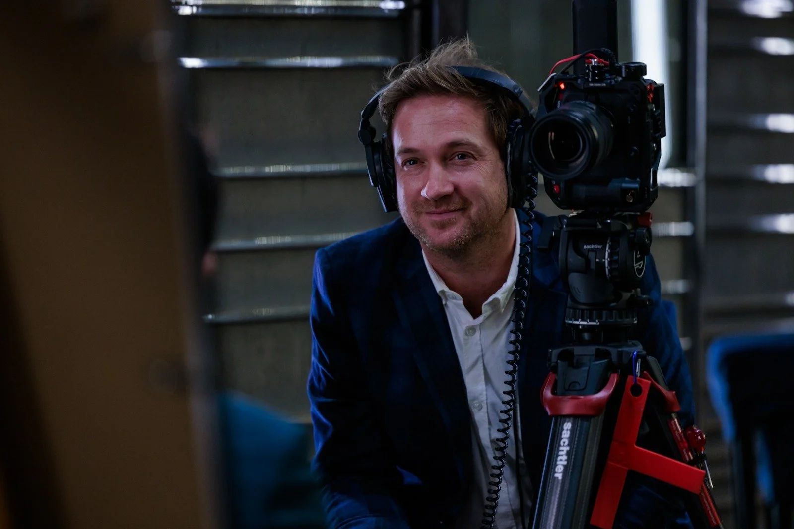 A man with light brown hair and a beard wearing a white shirt and a dark blazer, smiling, seated near a camera on a tripod with a camera operator headset.