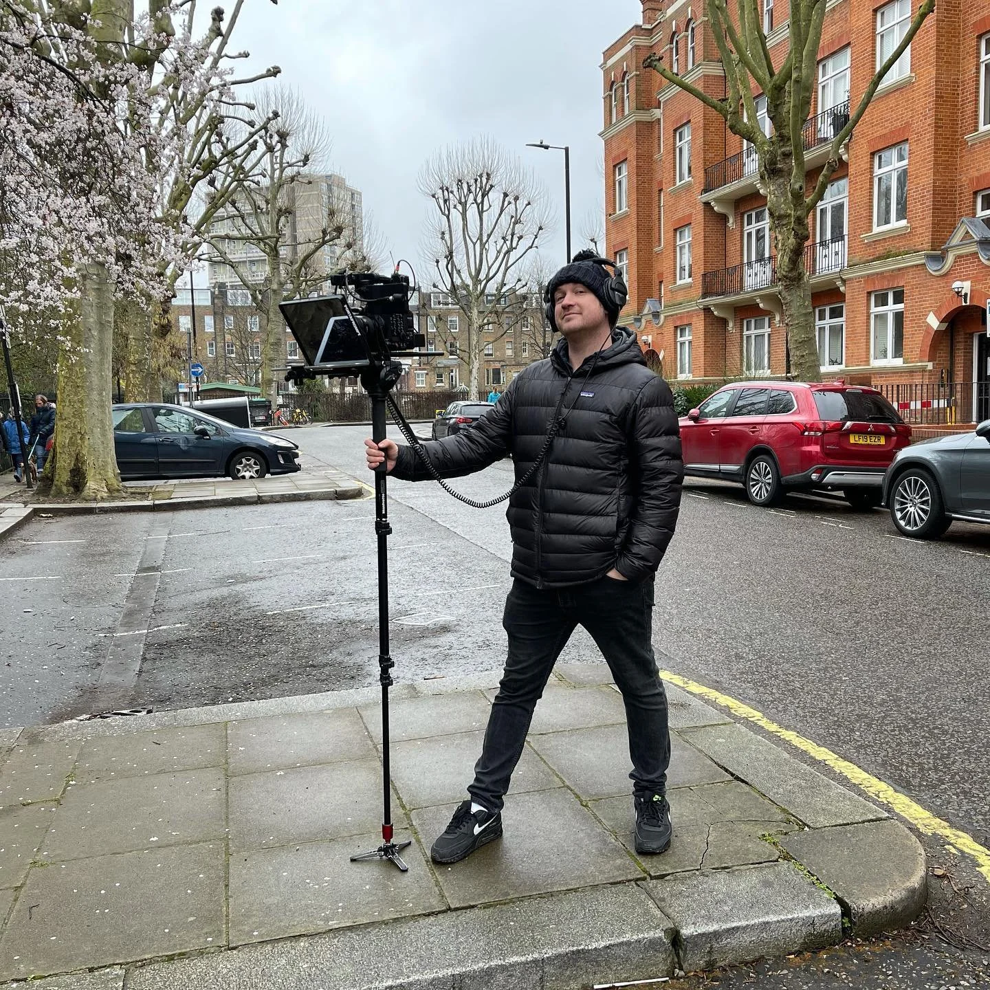 A man in a black puffer jacket, black pants, and sneakers standing on a sidewalk, holding a camera mounted on a tripod, with a street and apartment buildings in the background.