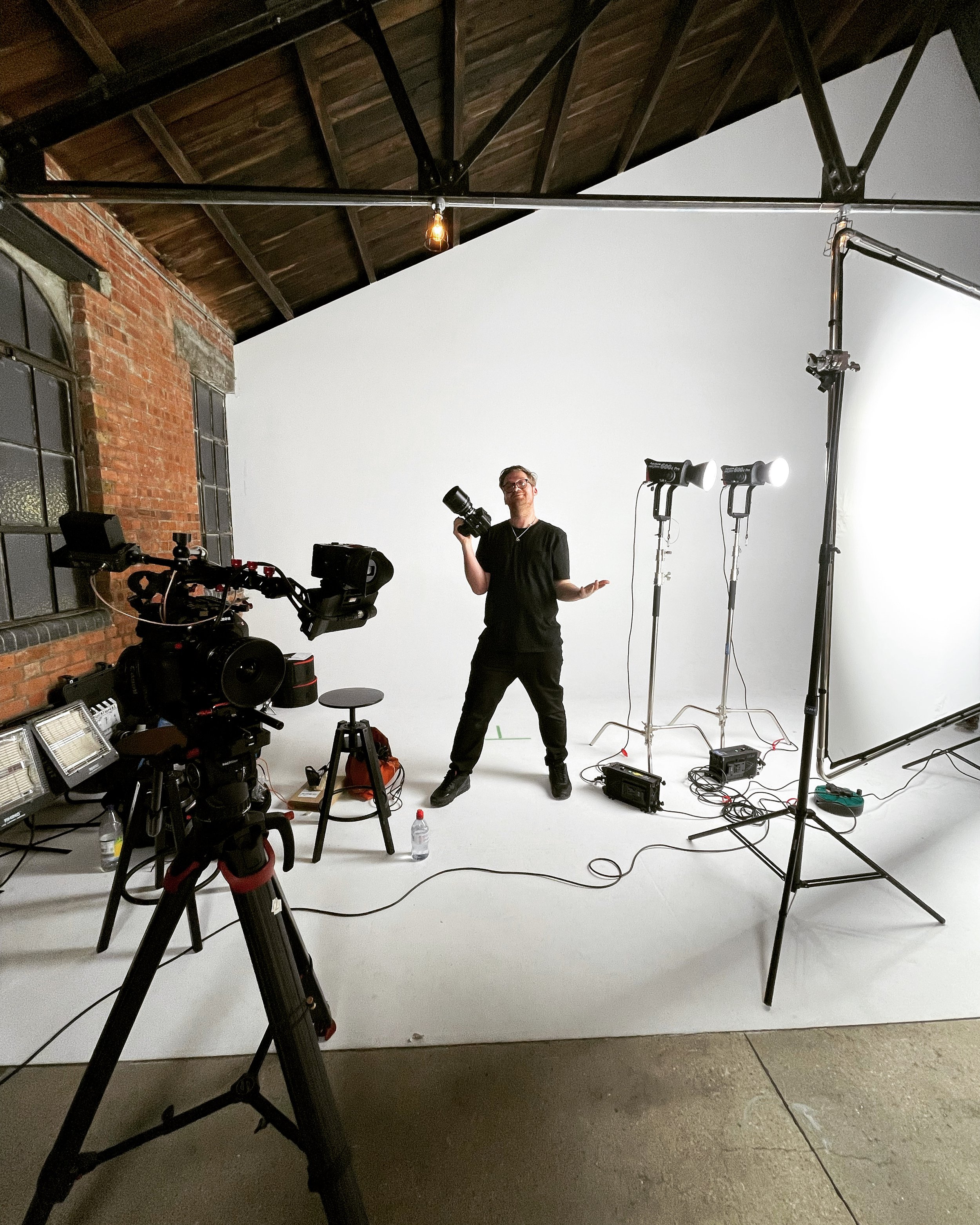 A photographer in a studio with a white backdrop, holding a camera, surrounded by lighting equipment and camera gear, in an industrial-style space with exposed brick walls and a wooden ceiling.