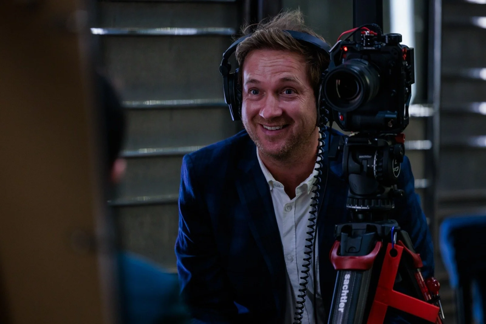 A man with a beard and brown hair smiling while wearing headphones, operating a professional video camera mounted on a tripod.
