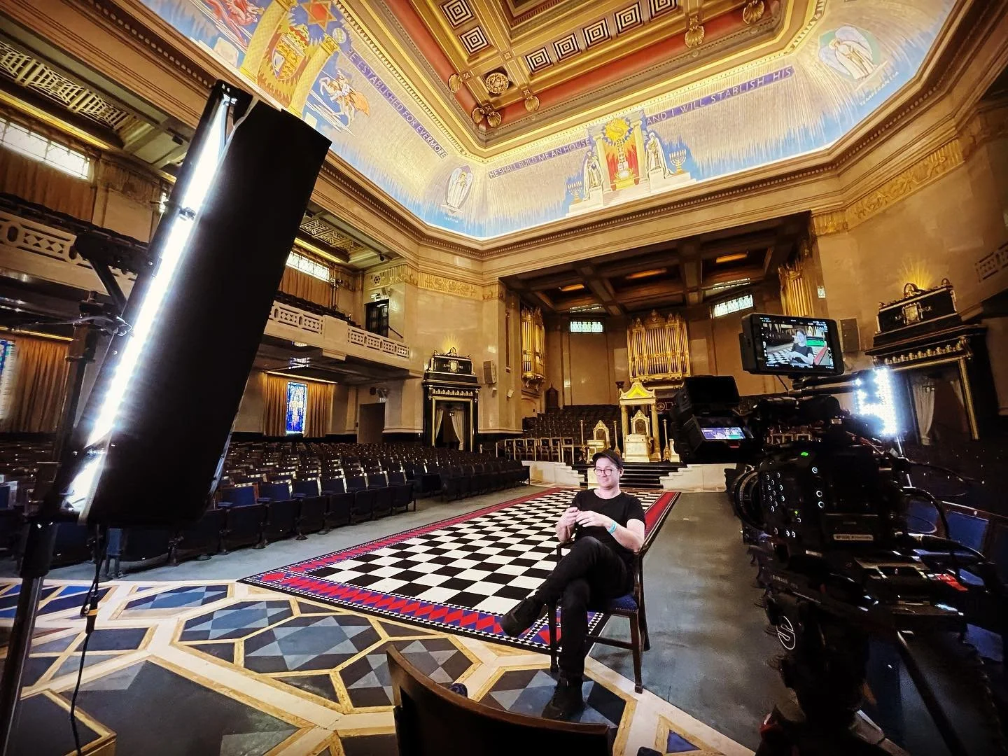 A person sitting in a large ornate hall on a checkered floor, surrounded by filming equipment.