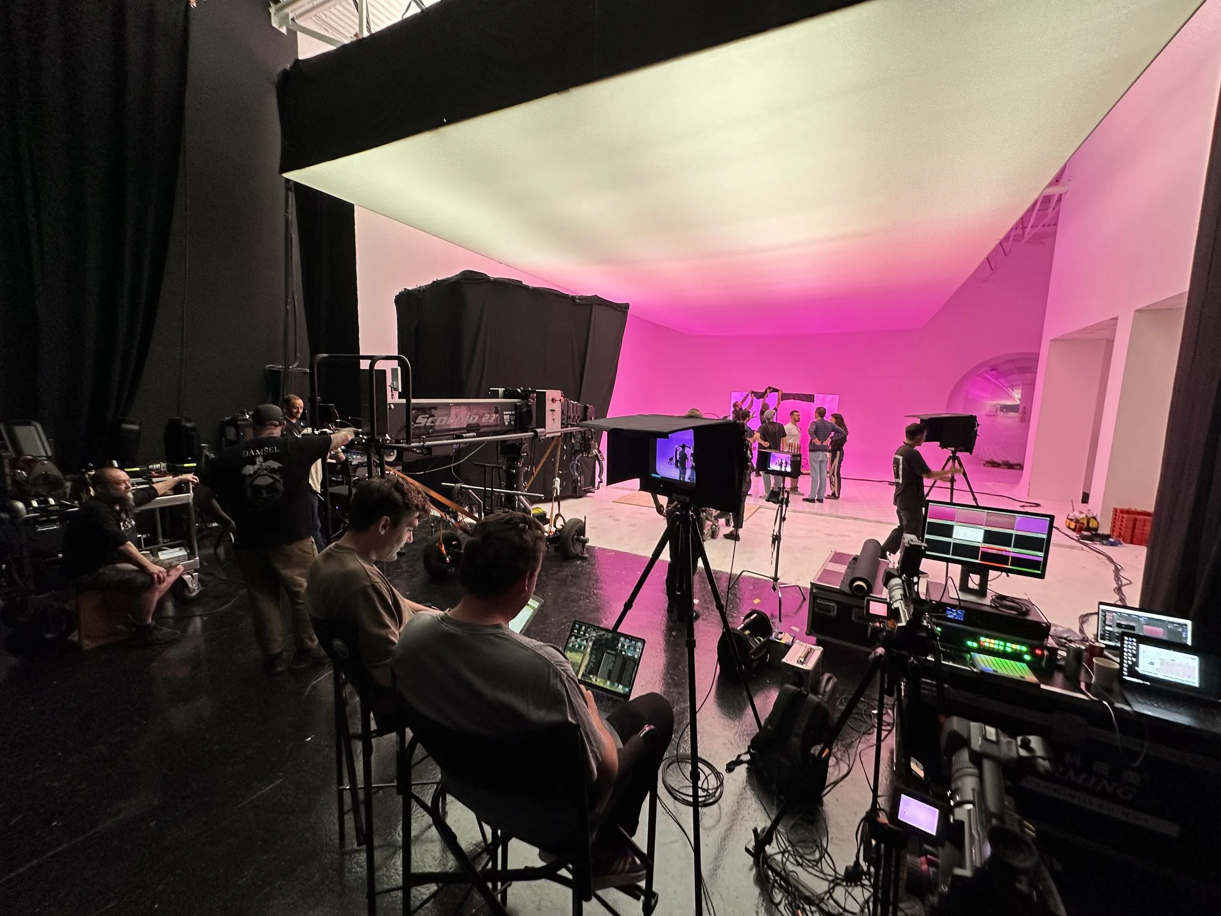 Behind the scenes of a film or photoshoot studio with crew members, equipment, cameras, and a large pink and white curved backdrop, with some individuals standing and others working with technology.