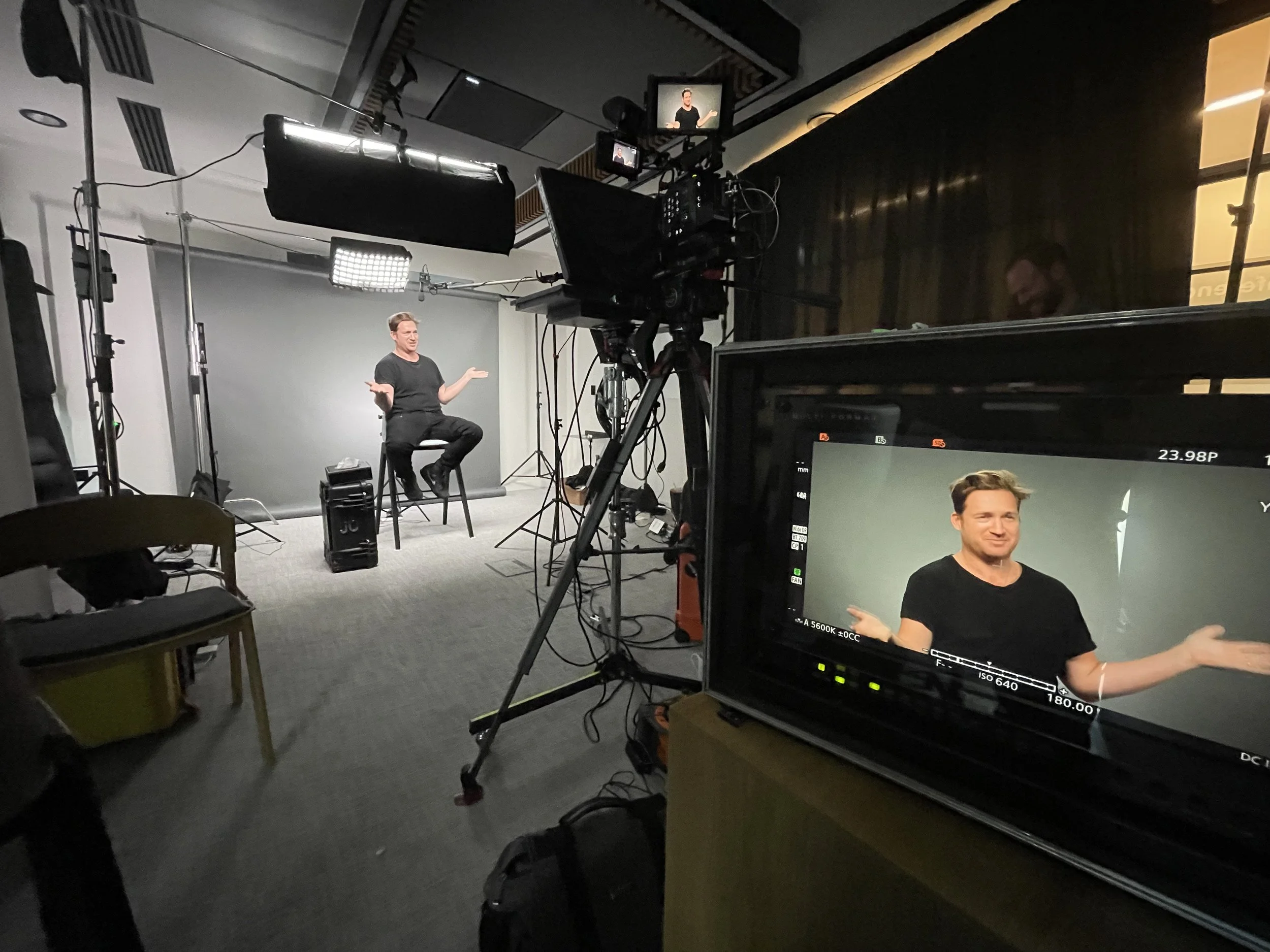 A behind-the-scenes view of a video shoot with a woman sitting on a chair and gesturing with her hands while speaking. Professional lighting and camera equipment are set up to film her, with monitor screens displaying her image. The setting appears to be a studio with a plain background.
