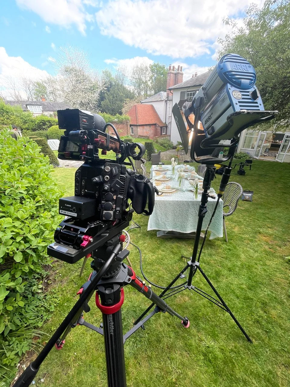 Professional camera and lighting equipment set up outdoors in a backyard with a dining table, chairs, and a garden, under a partly cloudy sky.