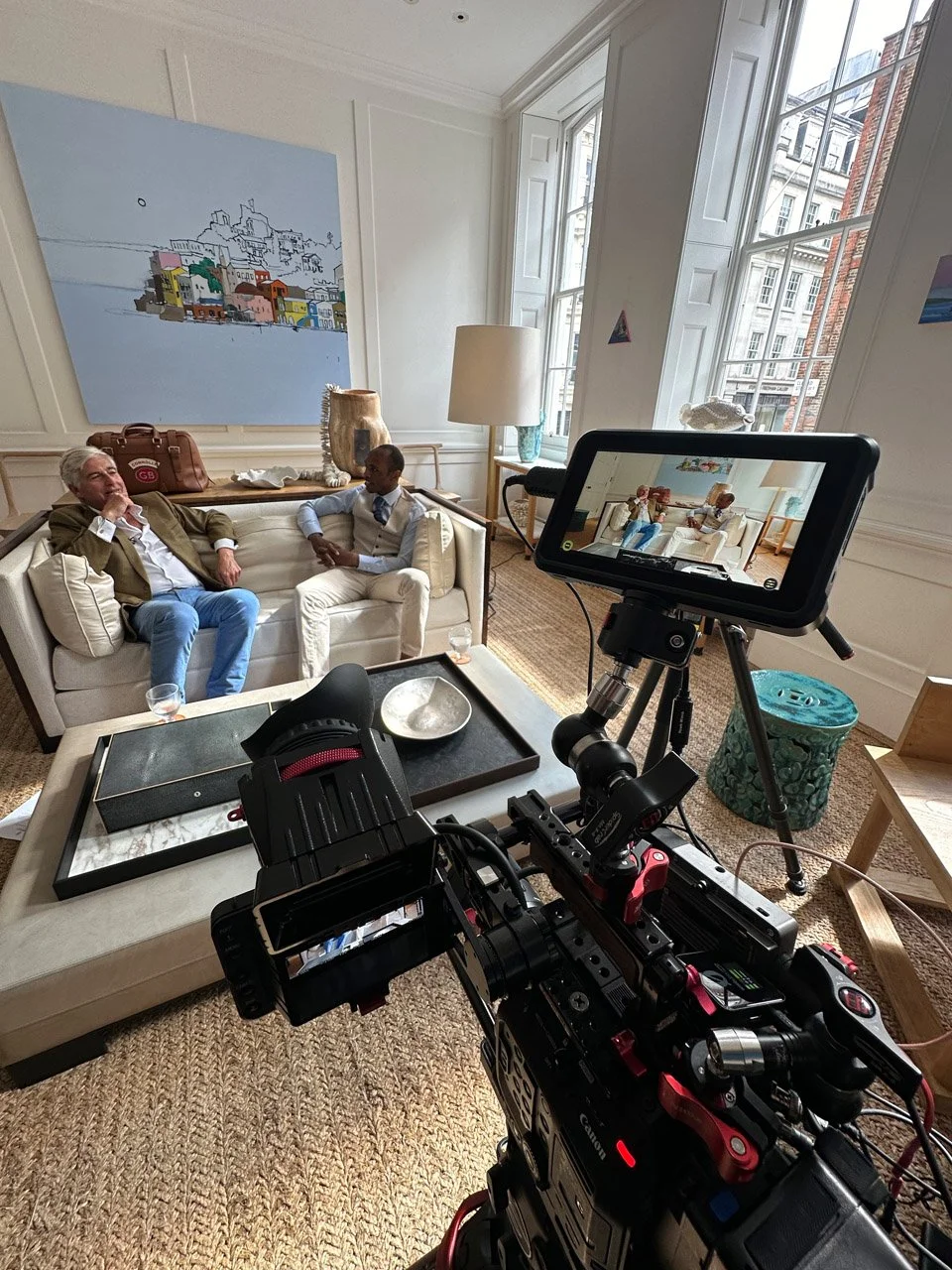Two men are sitting on a white sofa in a well-lit room with large windows. One man is dressed in a brown blazer and jeans, while the other wears a vest and dress shirt. A camera setup on a tripod is capturing the scene, with a monitor showing the view of the men. Artwork and a lamp are on the walls, and a table with decorative items is in the foreground.