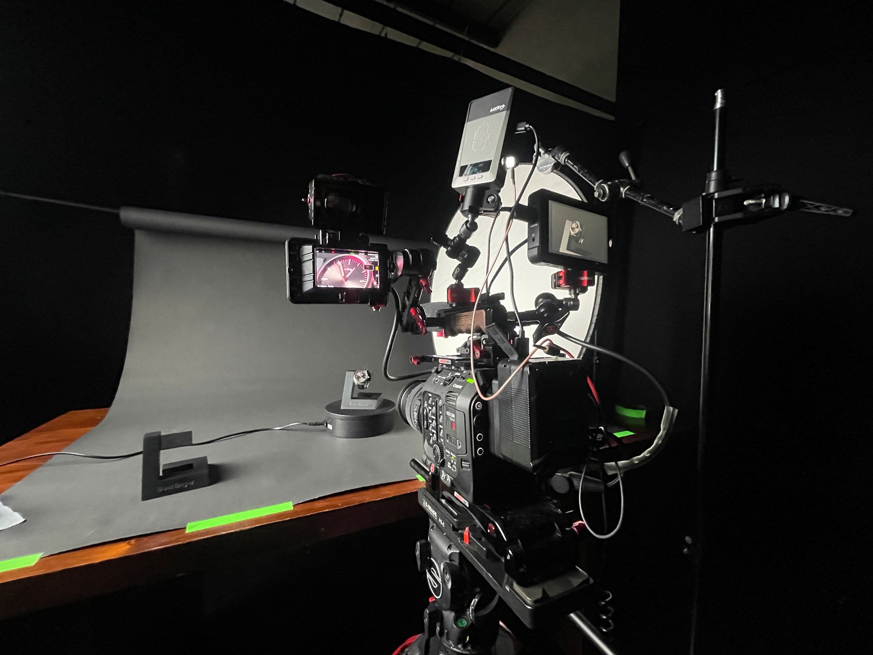 A professional camera setup with multiple monitors and a camera mounted on a tripod, positioned in front of a grey backdrop on a wooden table.