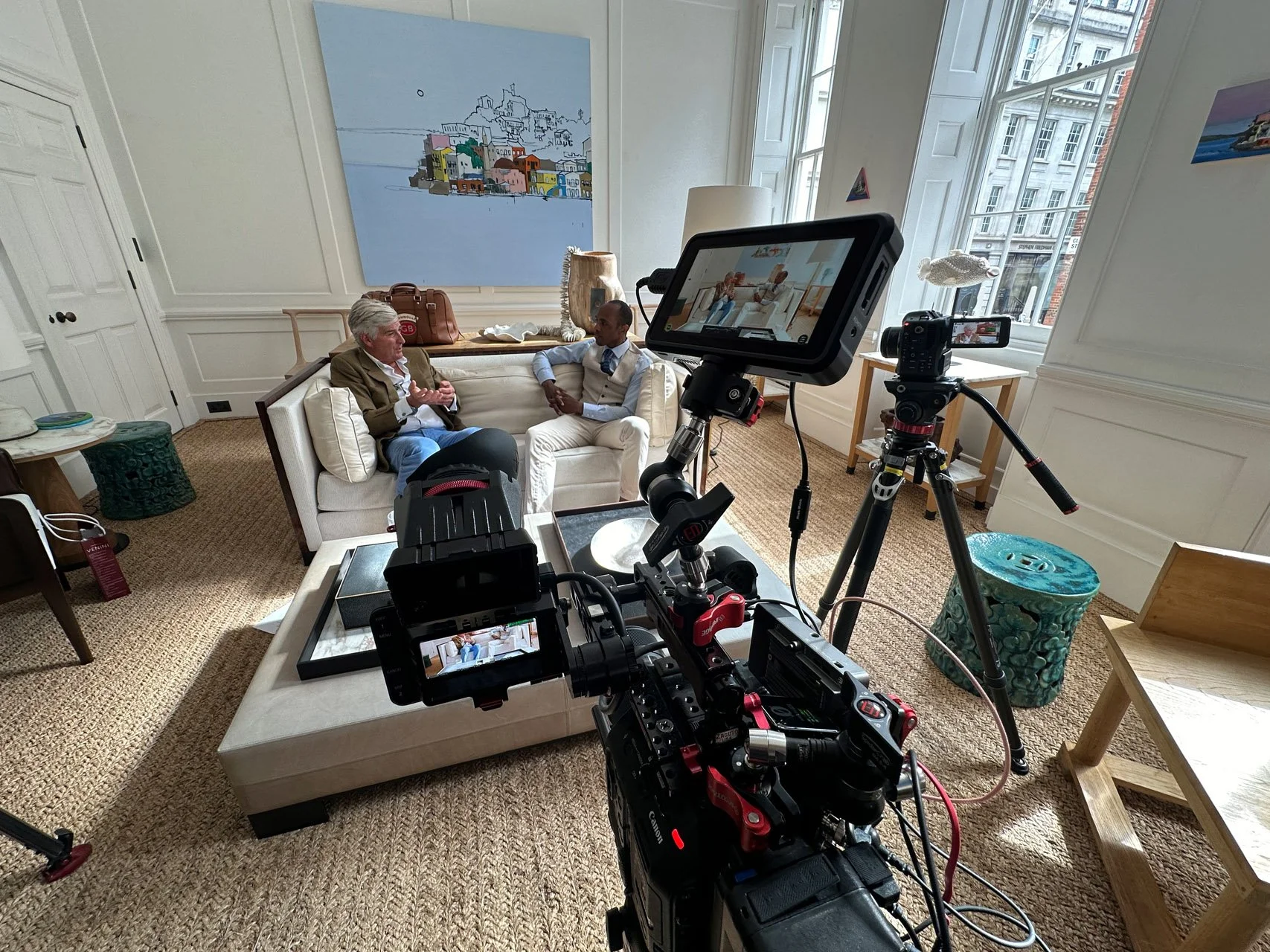 Two men are seated on a white sofa in a brightly lit room with large windows, having a conversation as part of an interview or recording session. Camera equipment and monitors are set up in the foreground. The room has artwork on the walls and various furniture pieces.