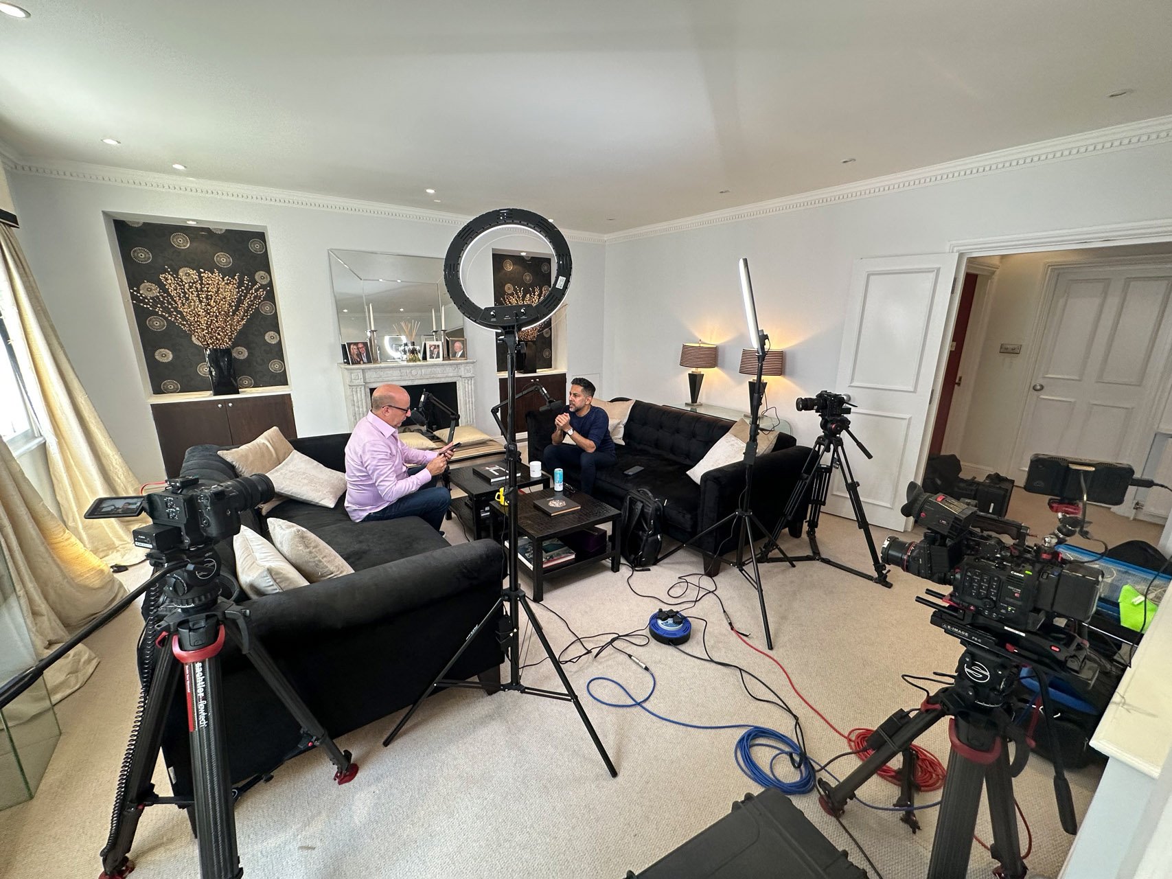 Two men are seated on black sofas in a large living room set up for filming, surrounded by professional camera equipment and lighting. One man is wearing a pink shirt and glasses, while the other is dressed in a dark blue shirt. A mirror above a fireplace reflects the scene, and there are decorative elements like lamps and artwork in the background.