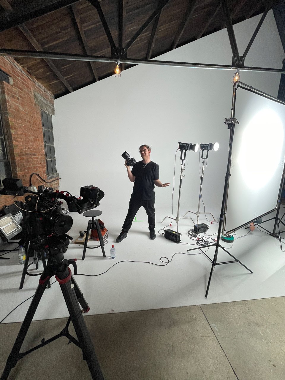 A man standing in a photography studio holding a camera, surrounded by professional lighting equipment, a white backdrop, and a camera on a tripod, with brick and metal wall features and a wooden ceiling.