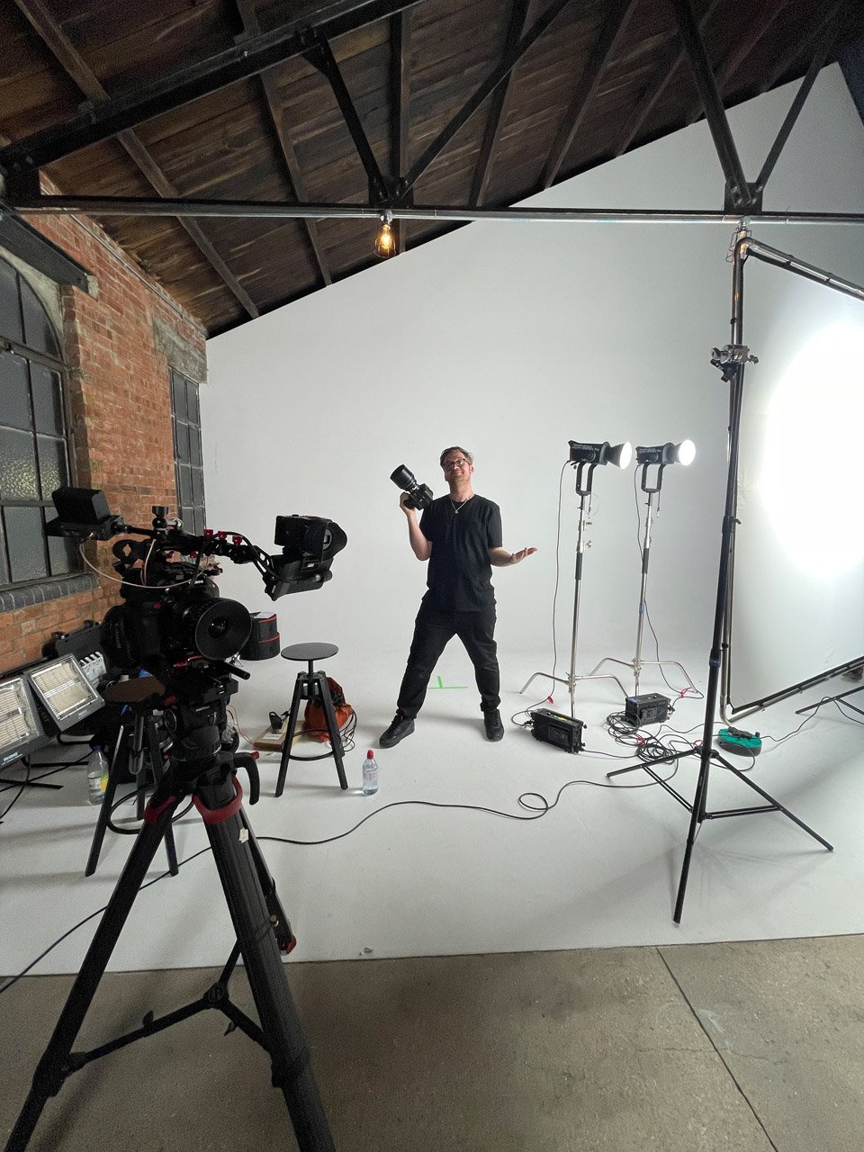 A person stands in front of a white backdrop in a studio, holding a camera, surrounded by professional lighting and camera equipment.