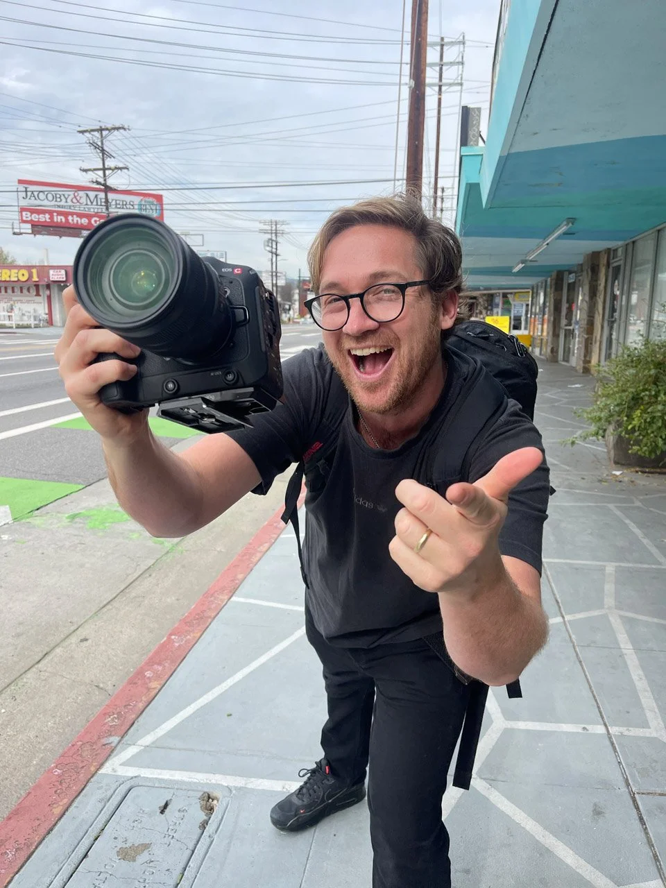 A man with glasses and a beard, smiling and pointing at the camera, holds a large camera lens in his hand on a city sidewalk.