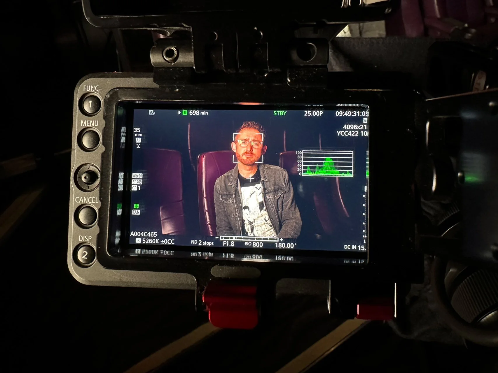 A camera monitor displaying a man with glasses and a denim jacket, sitting in a theater or cinema seat with a dark background.