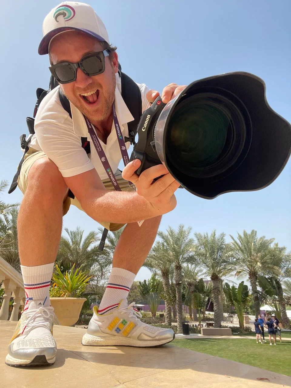 A man crouching outdoors, holding a professional camera and smiling at the camera. He is wearing sunglasses, a white cap, white sneakers with colorful details, and sports socks. Palm trees and a few people in the background suggest a warm, sunny environment.