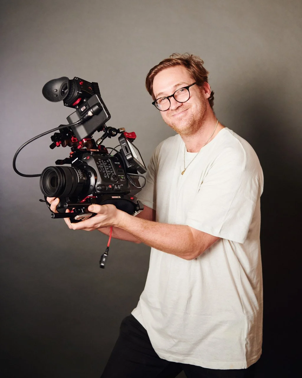 Man with glasses and a white t-shirt holding a professional video camera, smiling at the camera, against a plain gray background.